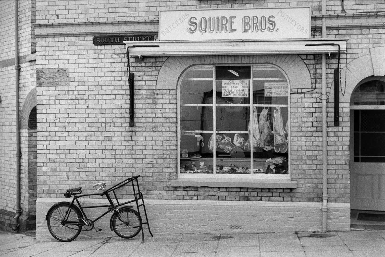 The shop front of Squire Brother's Butchers shop, in South Street, Torrington. A bicycle is parked outside and cuts of meat are on display in the window.