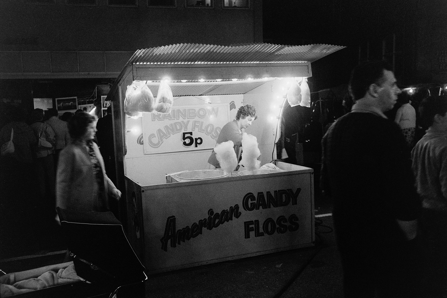 A woman selling American candy floss from a kiosk, at night in Northam, possibly at a fair or carnival. Men and women are walking past.