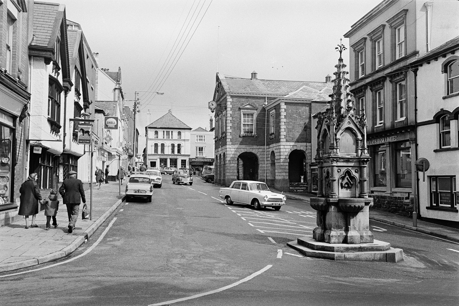 Street with shop fronts, parked cars and a water pump in Torrington. Shoppers are walking along the street and the Town Hall can be seen on the right with arches.