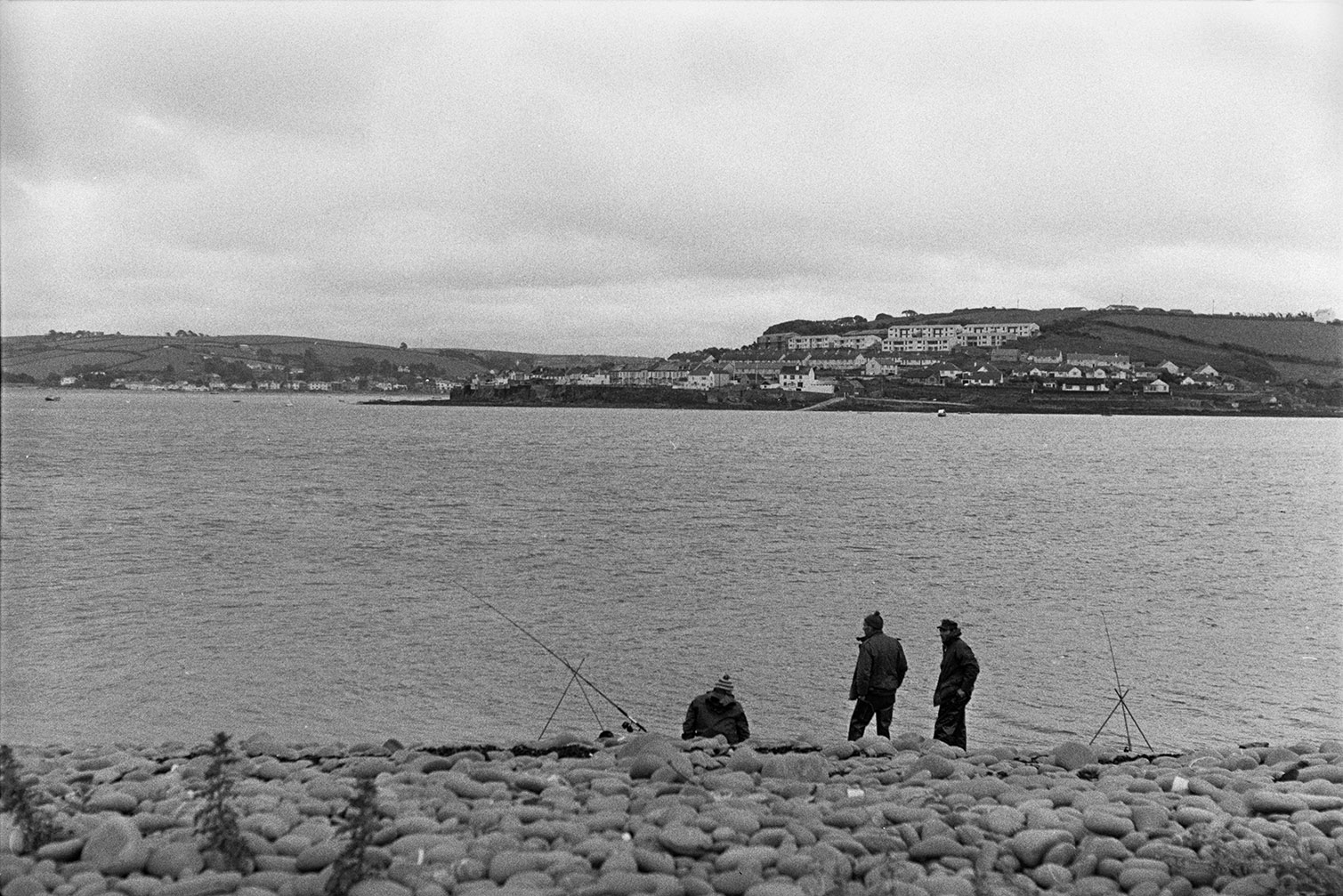 Three people fishing on a pebble beach with a town in the distance.