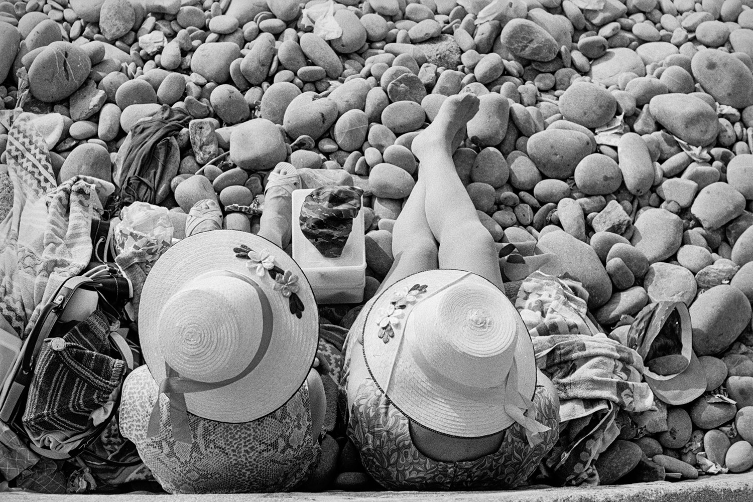 Two women, wearing sun hats, sat on a pebble beach with bags.