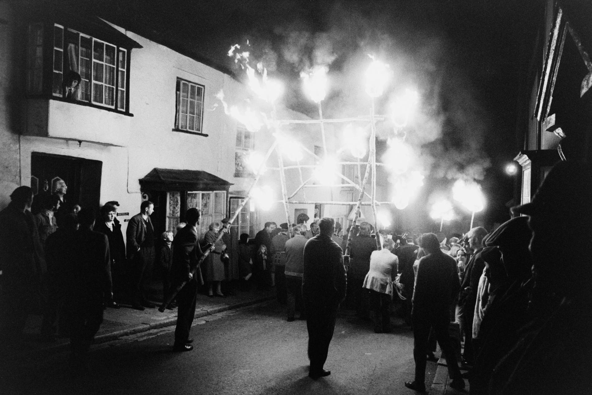 Spectators watching men carrying a wooden frame with flaming torches, in a torchlight procession through a street at Hatherleigh Carnival, at night.
