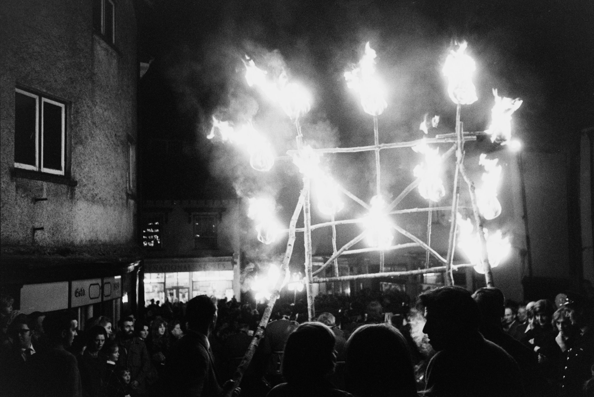 Spectators watching people carrying a wooden frame with flaming torches, in a torchlight procession through a street with shop fronts at Hatherleigh Carnival, at night.