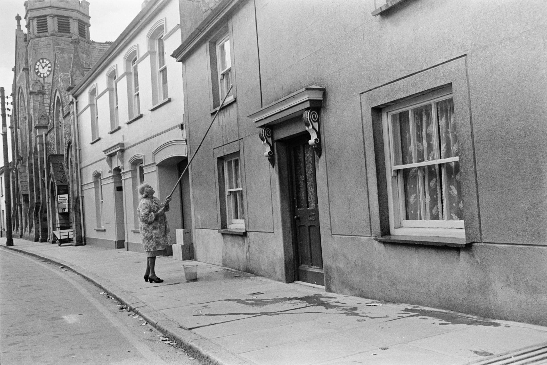 A woman cleaning first floor windows from the street outside, in Holsworthy. A church is visible in the background.