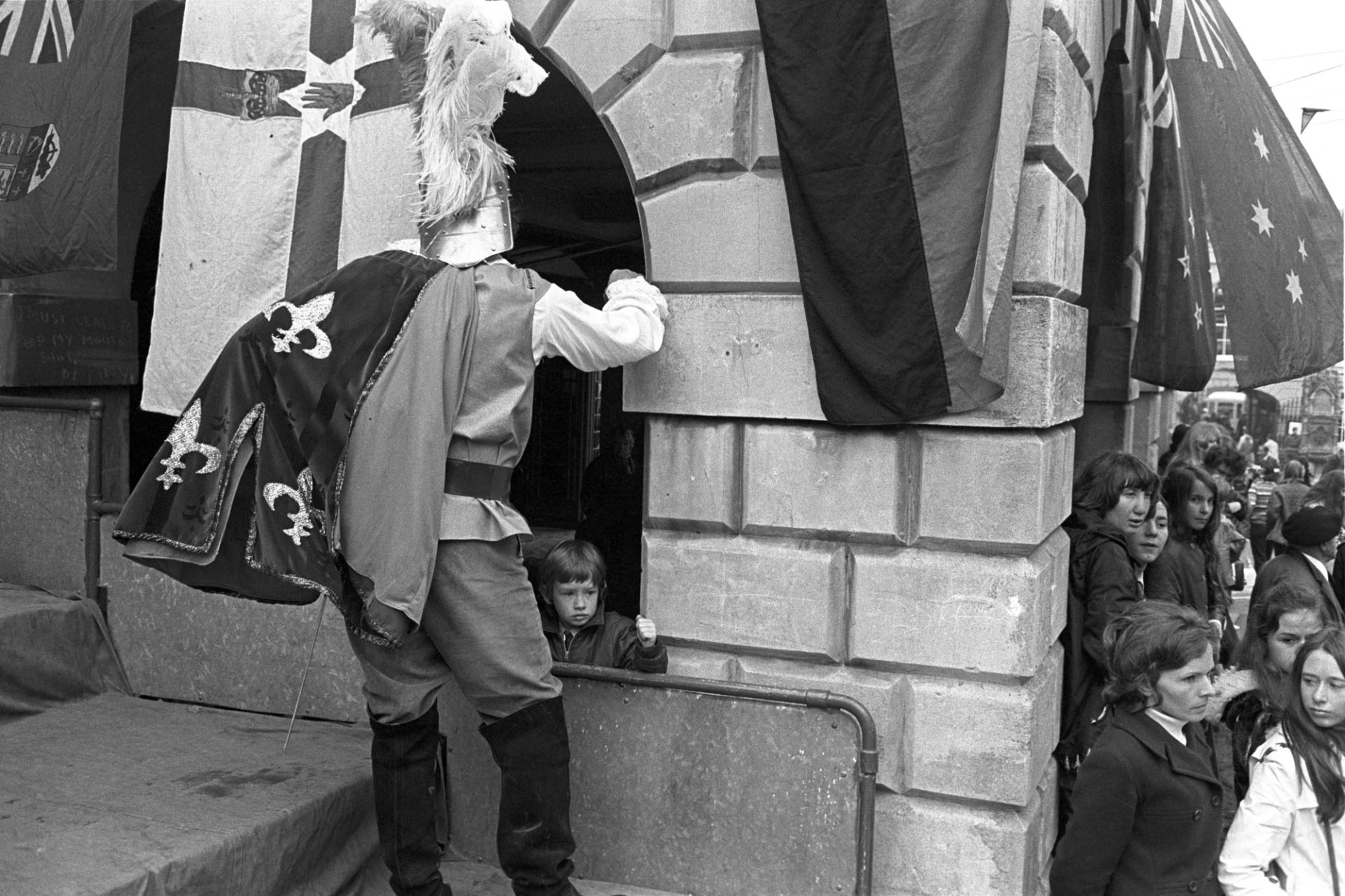 Torrington May Fair. Crowds and performers in High Street.
[People at Torrington May Fair in Torrington High Street. A person dressed as a knight looking into an archway partially covered by a flag, (possibly part of the Town Hall). A child is peering out of the archway.]