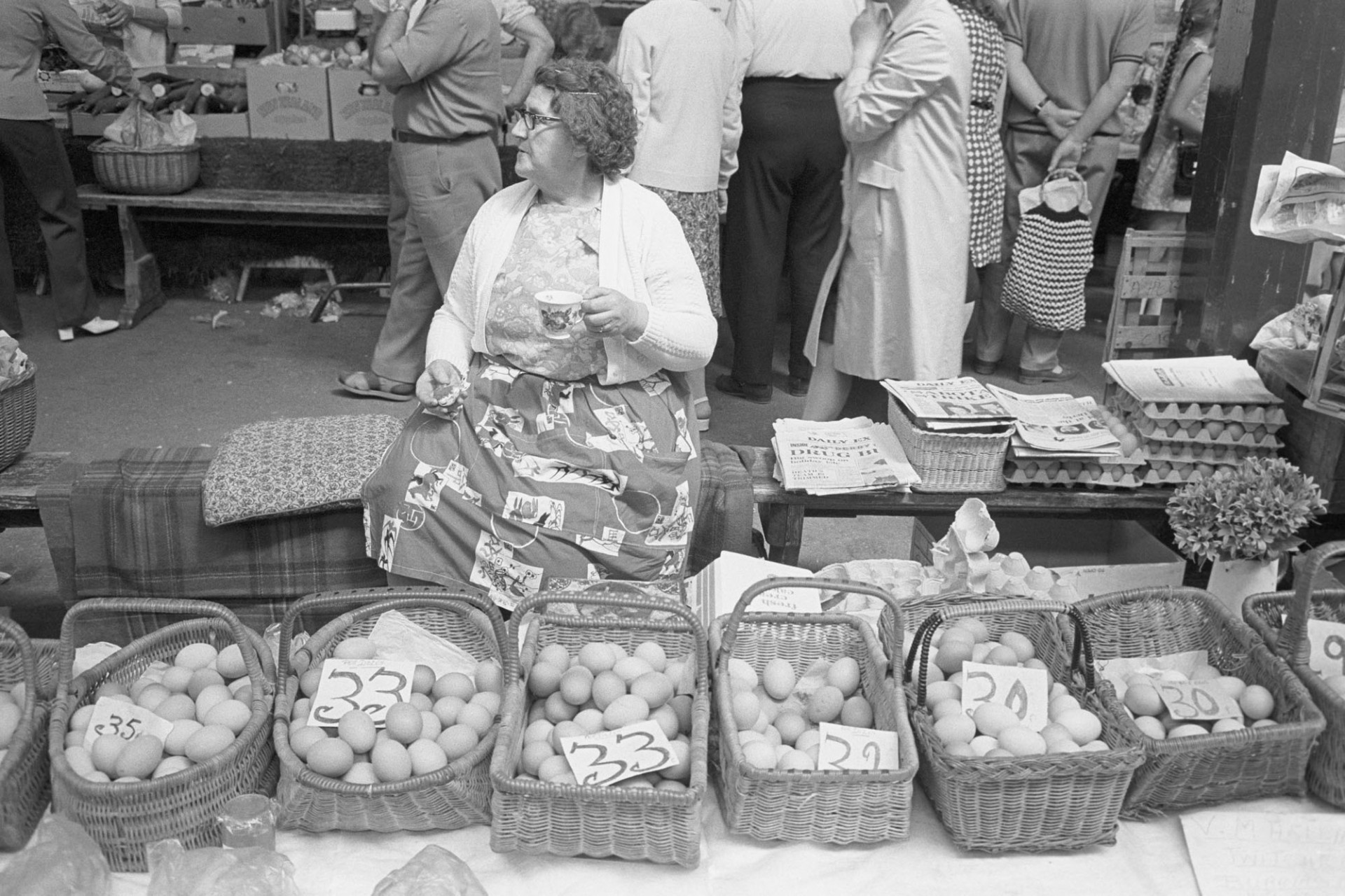 Mrs Pickard's poultry stall Pannier Market.
[Mrs Pickard drinking tea and selling eggs from wicker baskets at Barnstaple Pannier Market. The eggs are priced.]