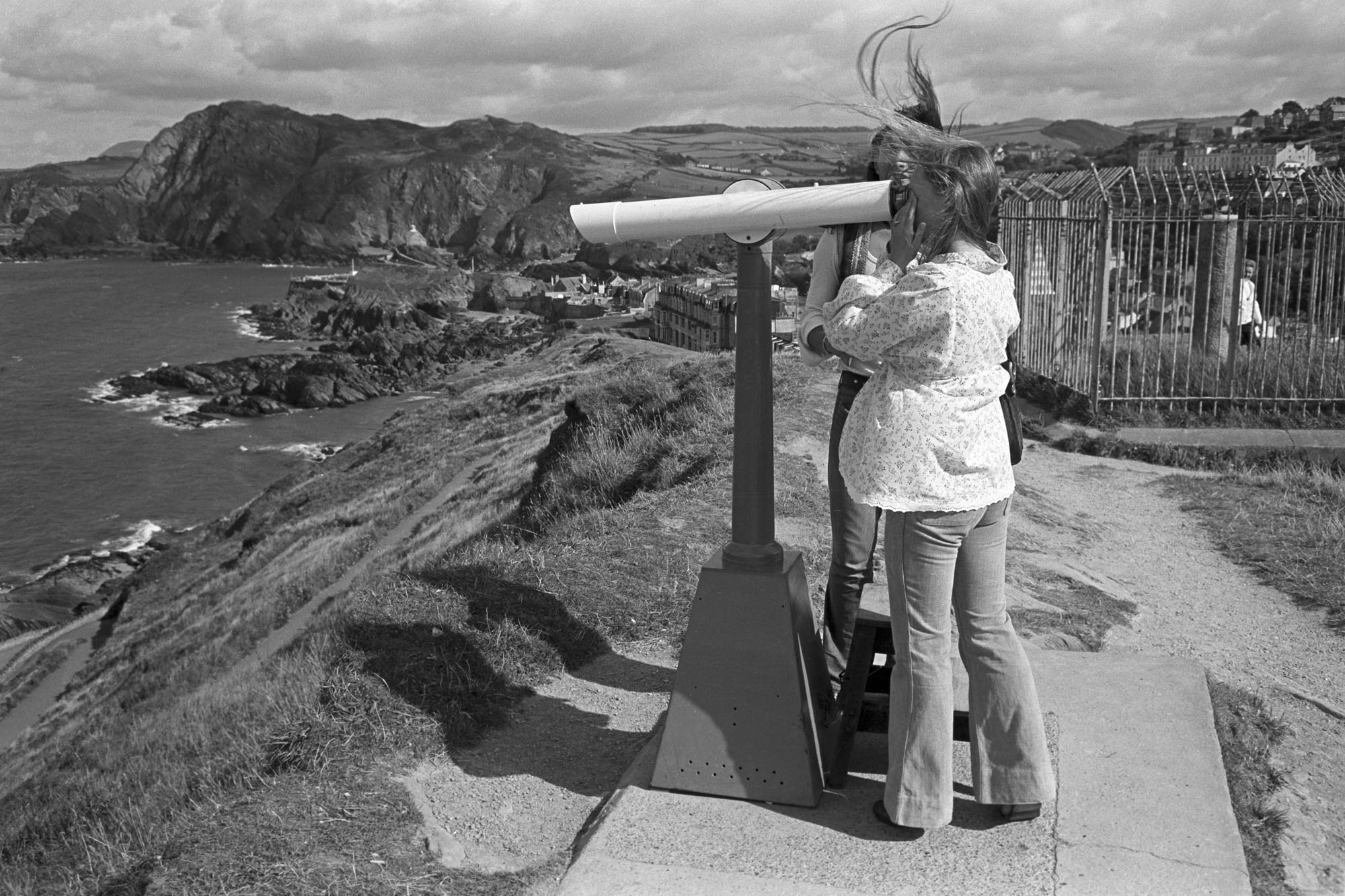 Couple looking through telescope, cliffs and sea.<br /> [A man and woman looking through a telescope on top of a cliff at the coastline below, at Ilfracombe.]