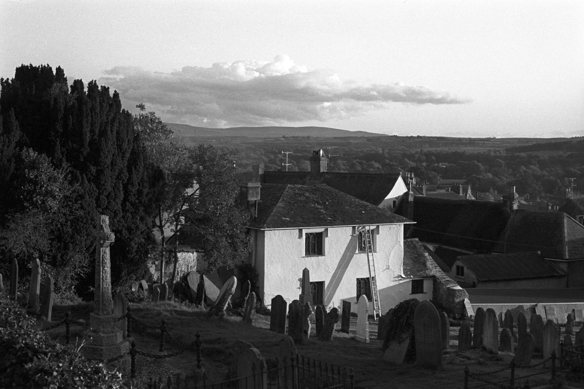 View over town towards Dartmoor, evening.
[Hatherleigh churchyard with gravestones, overlooking houses and a tree covered  landscape in the distance.]