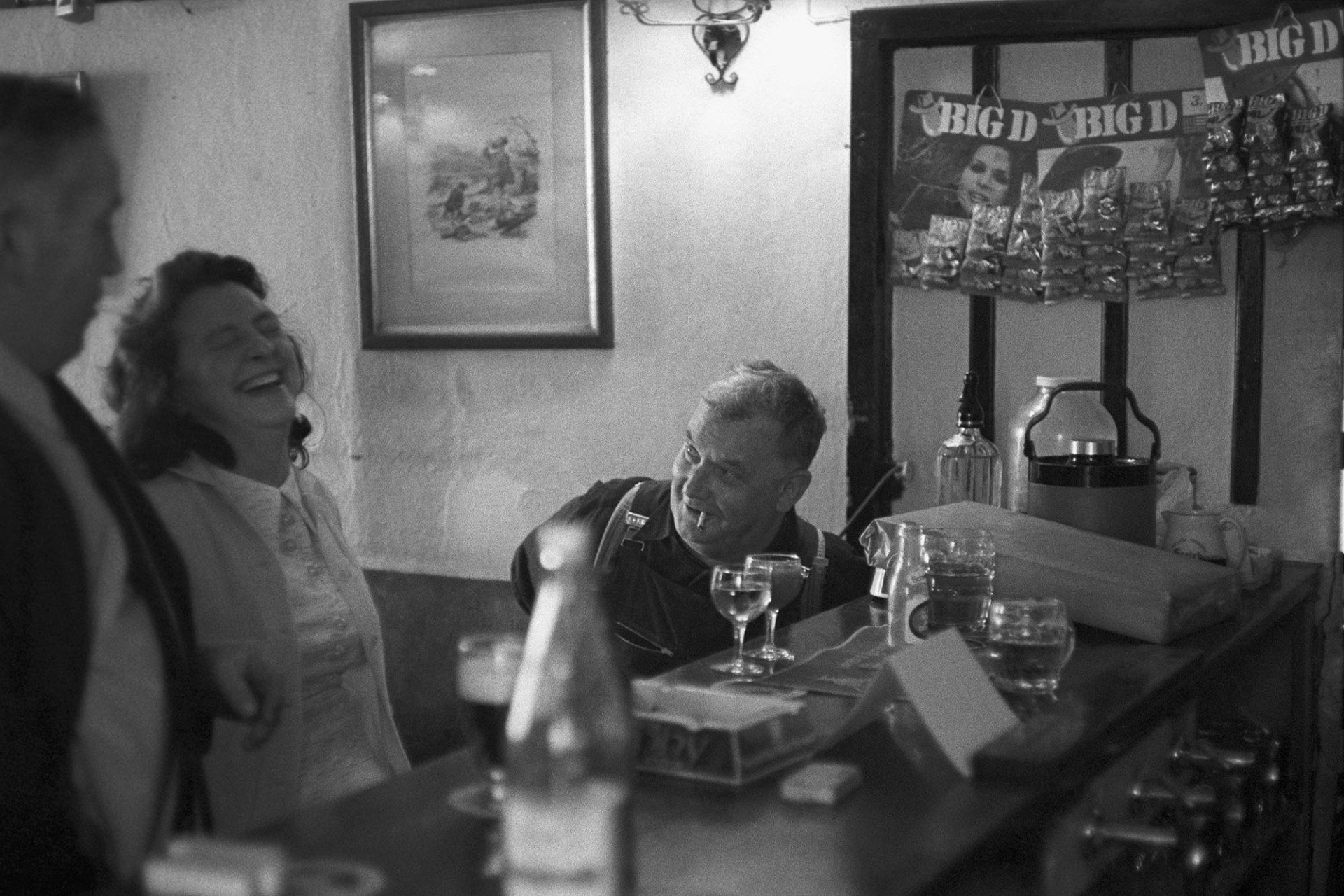 People enjoying a joke at the bar of The George.
[Two men and a woman laughing and drinking at the bar of The George in Hatherleigh. One man is smoking a cigarette and glasses of wine and a pint glass are on the bar.]
