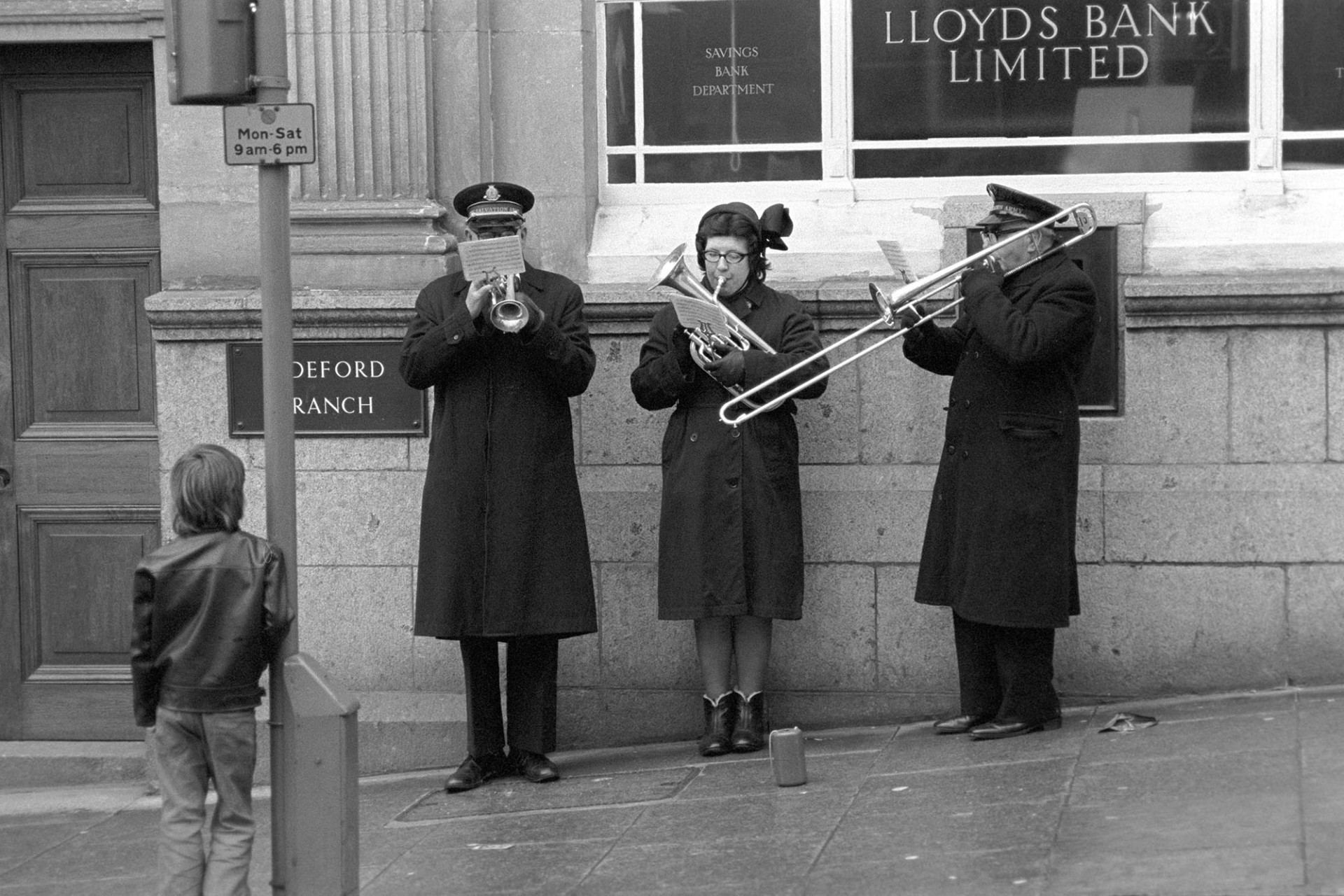 Salvation Army, three musicians playing to one child.
[Three musicians from the Salvation Army playing brass instruments, including a trombone, outside Lloyds Bank in Bideford High Street. A child is listening to them.]