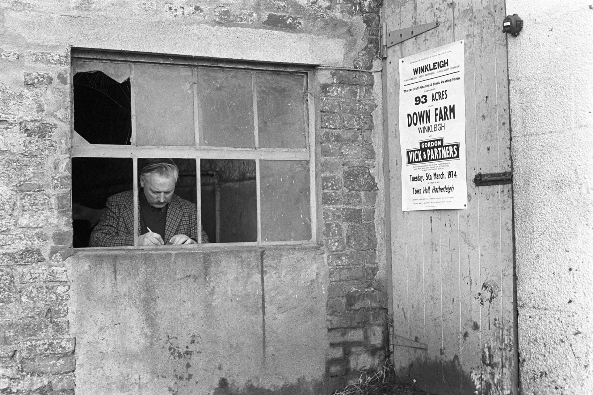 Auctioneers office at farm sale.
[A man sat by a window in the auctioneer's office at Hall Farm, Beaford. A poster advertising another sale is stuck to a door next to the window.]