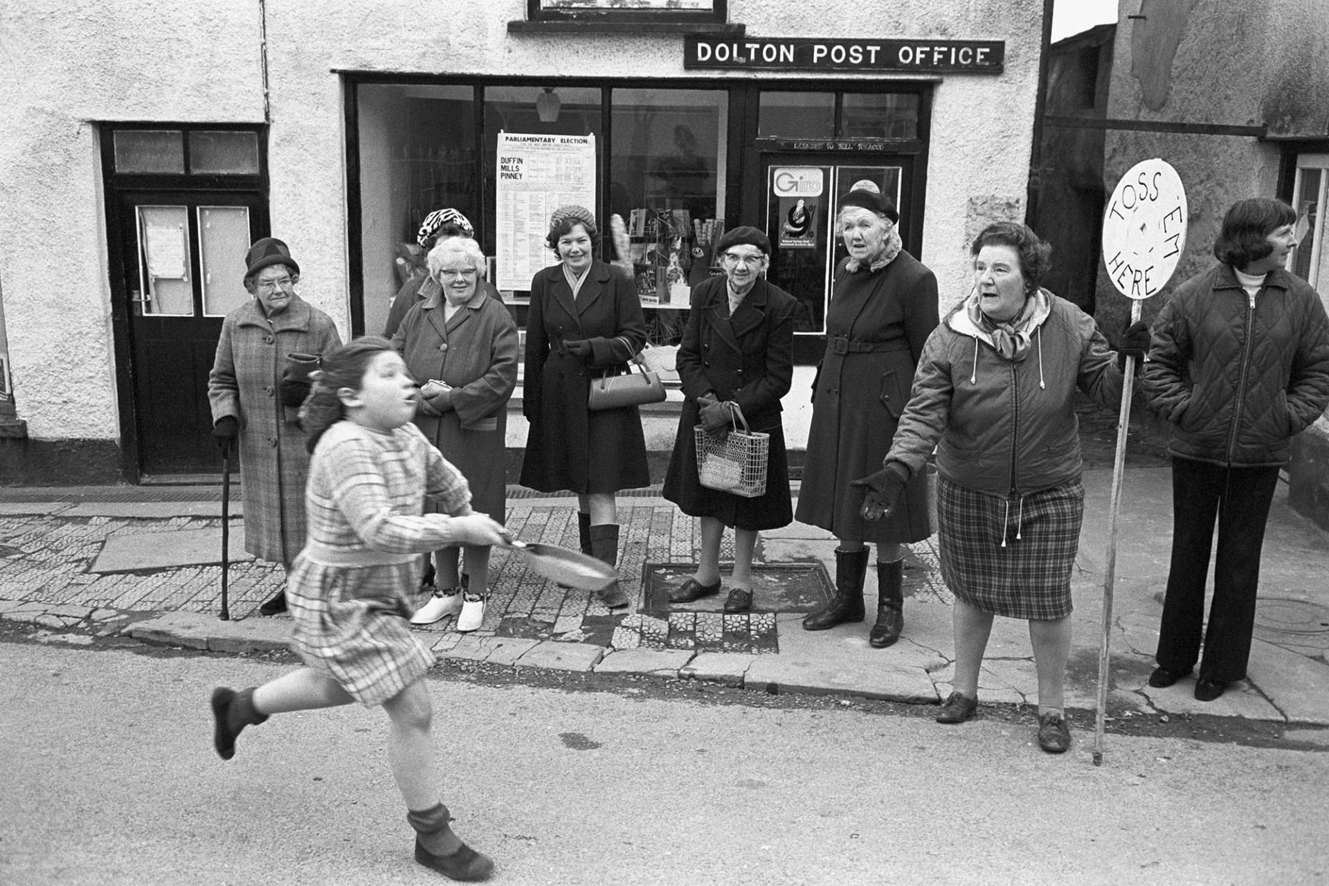Pancake race, runner and spectators outside Post Office.
[Spectators watching Diane Hiscock running in the pancake race outside Dolton Post Office. A woman with a 'Toss Em Here' sign is also outside the Post Office.]
