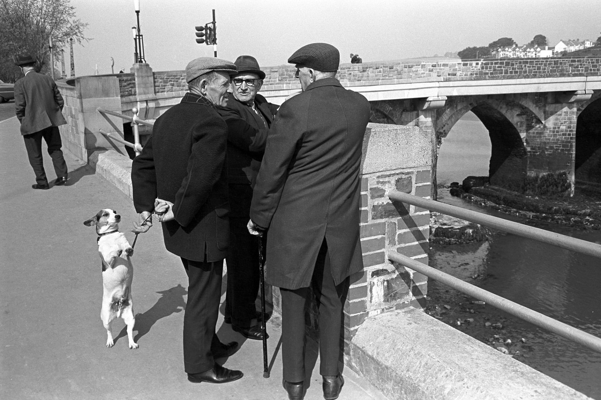 Three men chatting with dog, Humorous, Bridge behind. 
[Three men talking with a dog by Bideford Bridge. The bridge is also known as Bideford Long Bridge and Bideford Old Bridge.]
