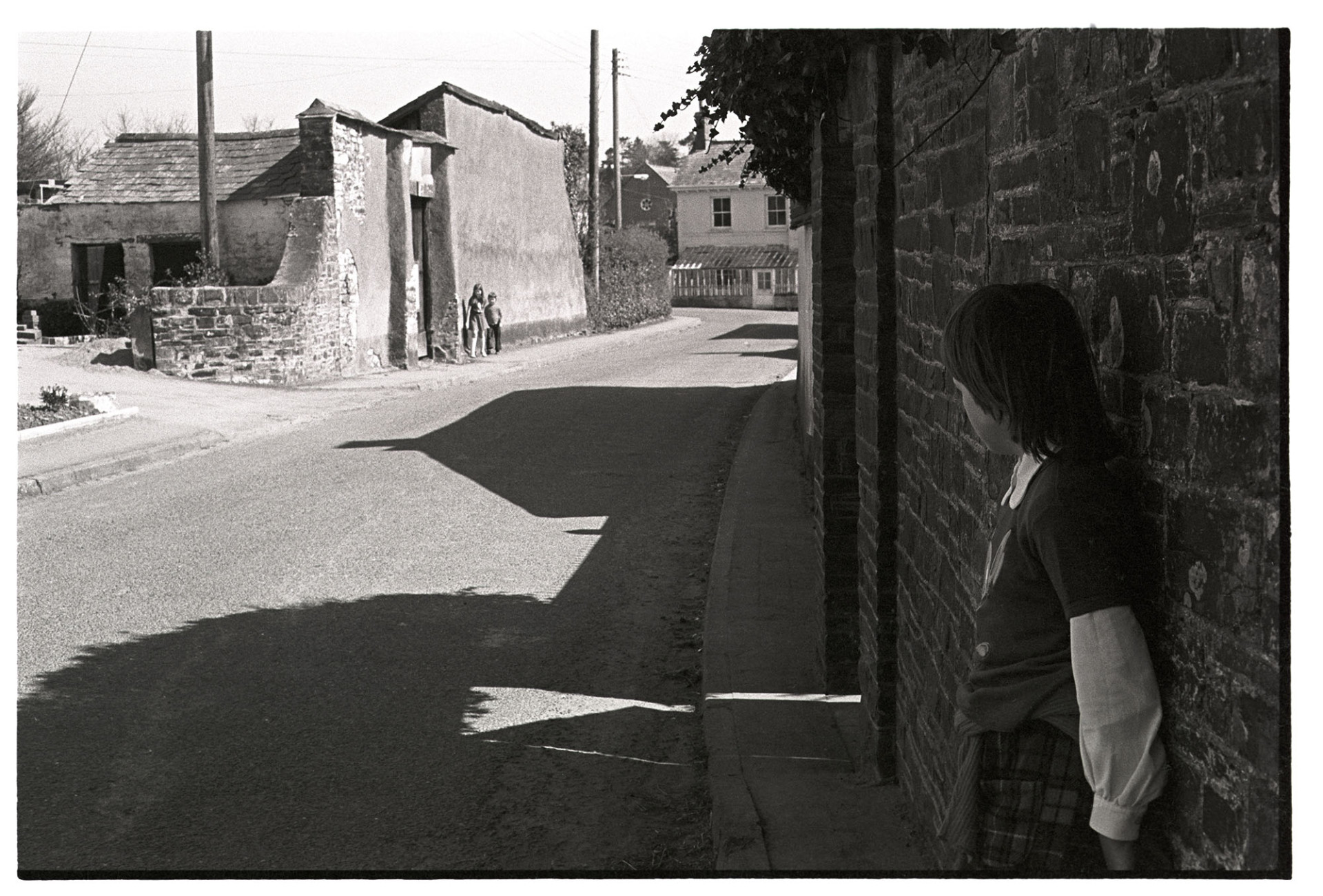 Village street with children. 
[Children playing in a street by a cob wall in Dolton. Shadows from buildings alongside the street and projected onto the road.]