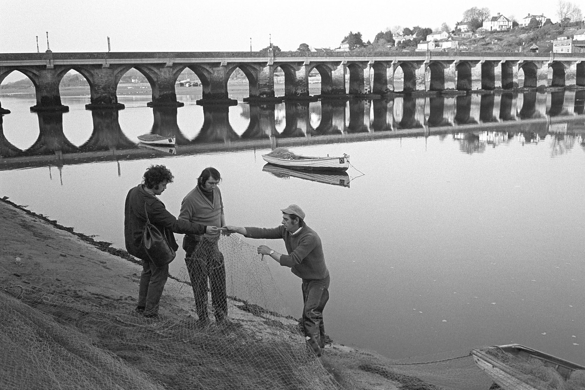 Salmon fishers mending nets in front of bridge. 
[Men checking their fishing nets on the banks of the river Torridge by Bideford Bridge. Rowing boats can be seen on the river. The bridge is also known as Bideford Long Bridge and Bideford Old Bridge.]