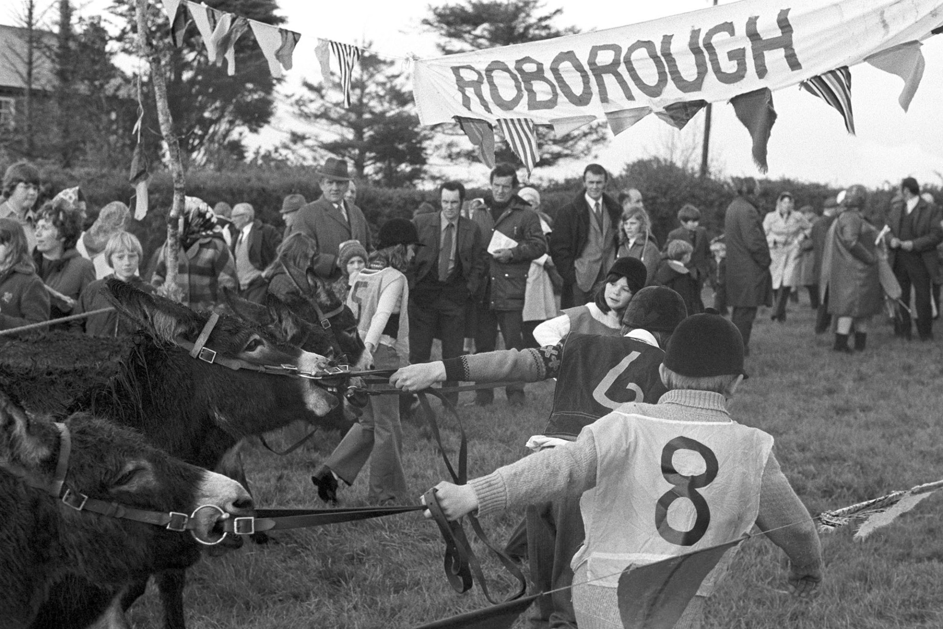 Donkey Derby, Jockeys and crowd. 
[Children leading their donkeys under a banner with 'Roborough' written on it, and past spectators watching the derby at Roborough.]