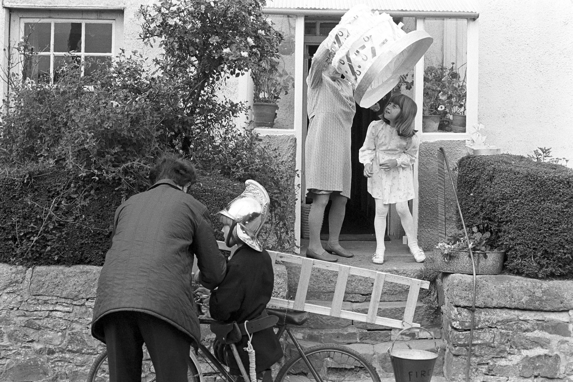 Children preparing for fancy dress parade. 
[Children outside their house getting ready in their costumes, with the help of a man and woman for a fancy dress parade at Winkleigh Fair. One child is dressed as a fireman and the other is dressed as a cake.]