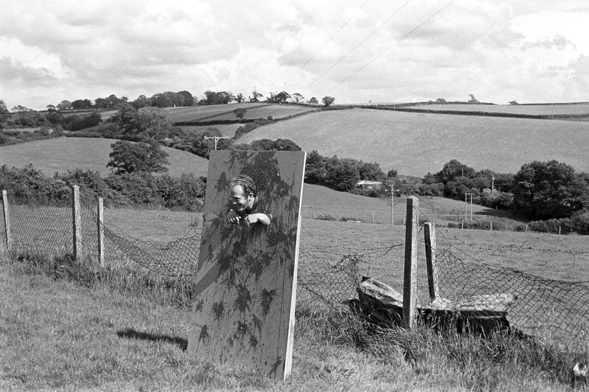 Man having tomatoes thrown at himself in stocks. 
[Man in stocks having tomatoes thrown at him at Chittlehampton pageant. A landscape of trees, field and hedgerows is in the background.]