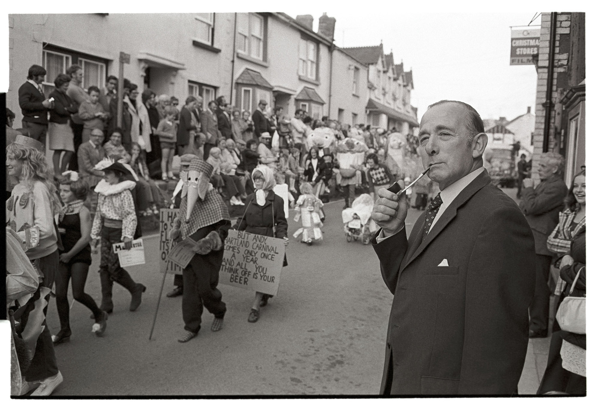 Man watching carnival parade smoking pipe.
[Man smoking a pipe watching people in fancy dress parade past at Hartland Carnival. Other spectators are watching the procession from the other side of the street.]