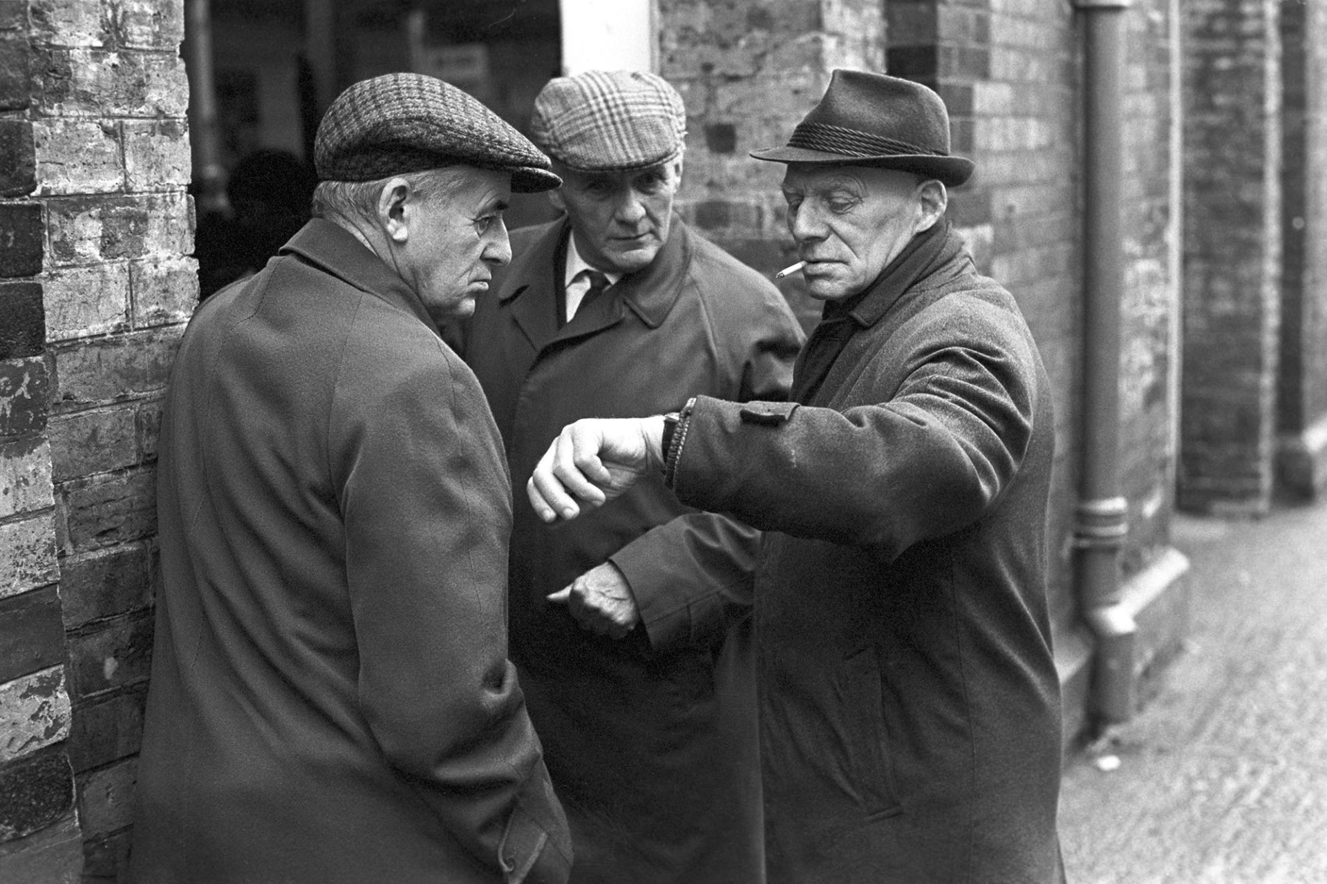 Three men chatting in street.
[Three men talking outside Barnstaple Pannier Market.  One of the men is smoking a cigarette and checking his watch.]