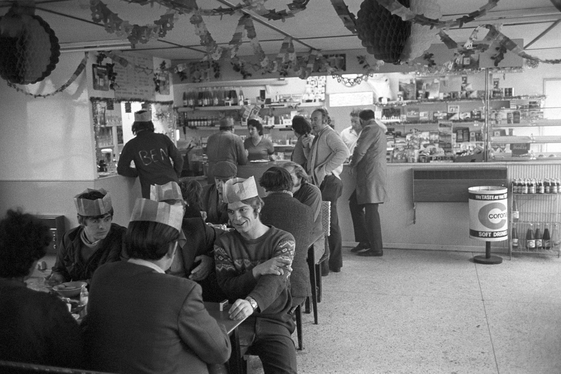 People in party hats having free Christmas Dinner.
[People, some wearing Christmas hats, sitting at tables and standing at the counter for Christmas dinner,  in the transport café at Winkleigh.]
