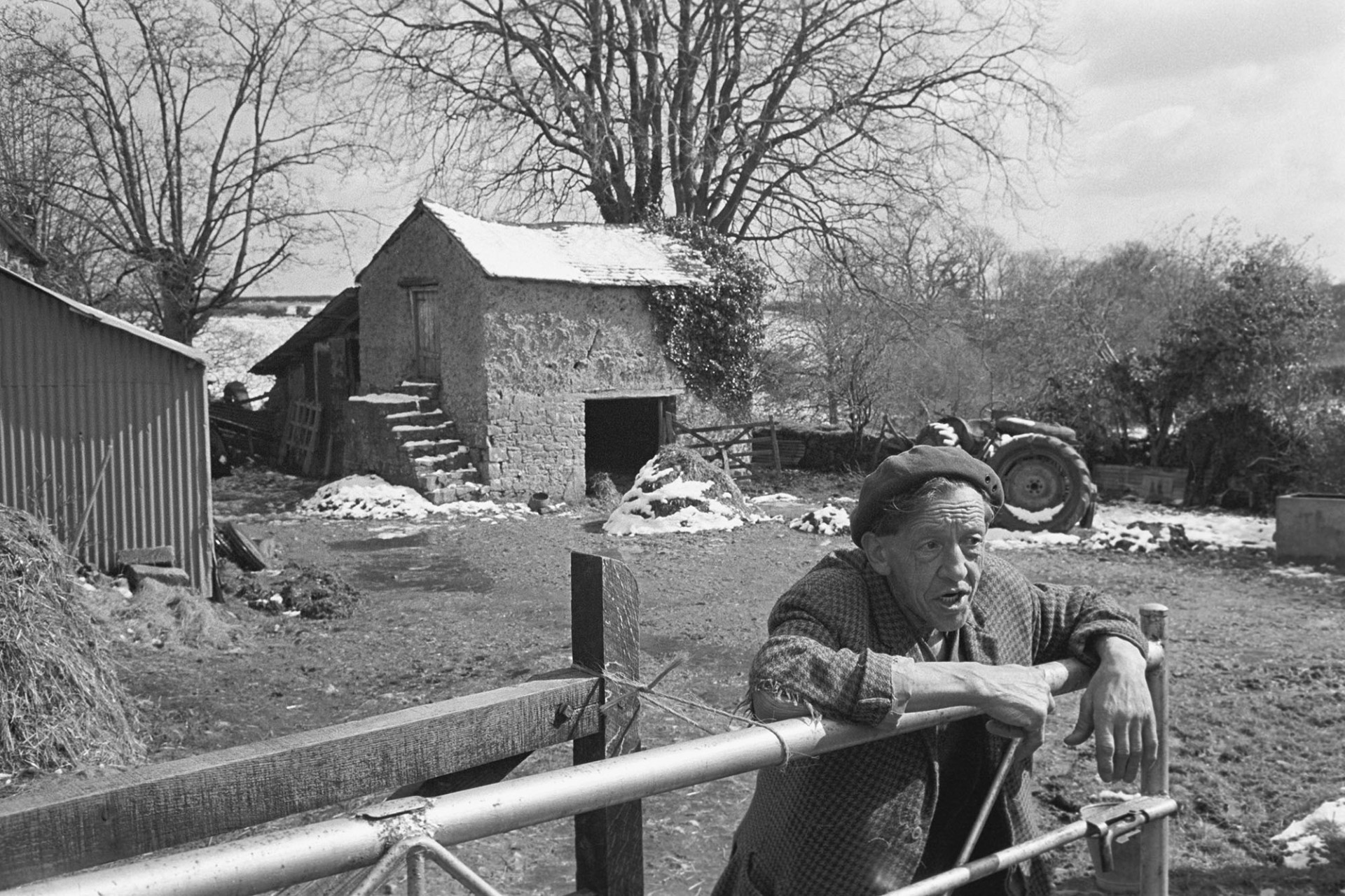 Farmer leaning over gate in farmyard, barn in background.
[Bill Cooke leaning over a gate in a farmyard. A corrugated iron barn, cob granary and muckheaps covered in snow are visible in the background, at Colehouse in Riddlecombe.]