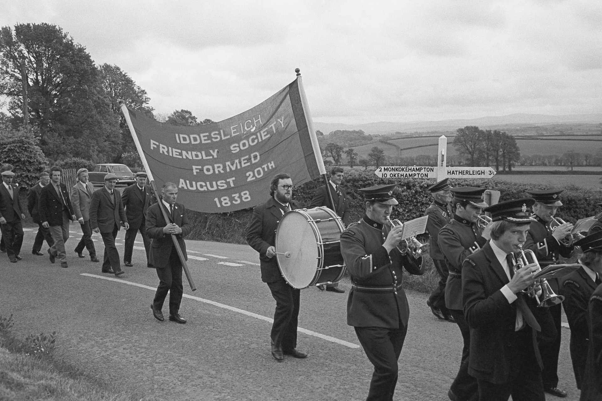Friendly Society Parade, silver band, banner and members.
[Hatherleigh Silver Band leading the Iddesleigh Friendly Society Club Day parade. Two men are carrying a banner in the parade. The parade is passing a signpost.]
