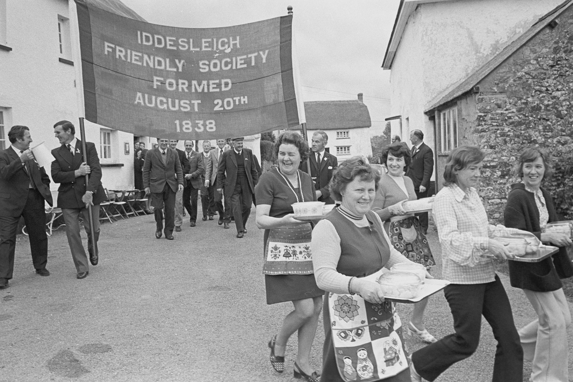 Friendly Society Members and bandsmen. Women parading the mashed potatoes to the lunch.
[Women carrying trays of food and men carrying an Iddesleigh Friendly Society banner in the Iddesleigh Club Day parade.]