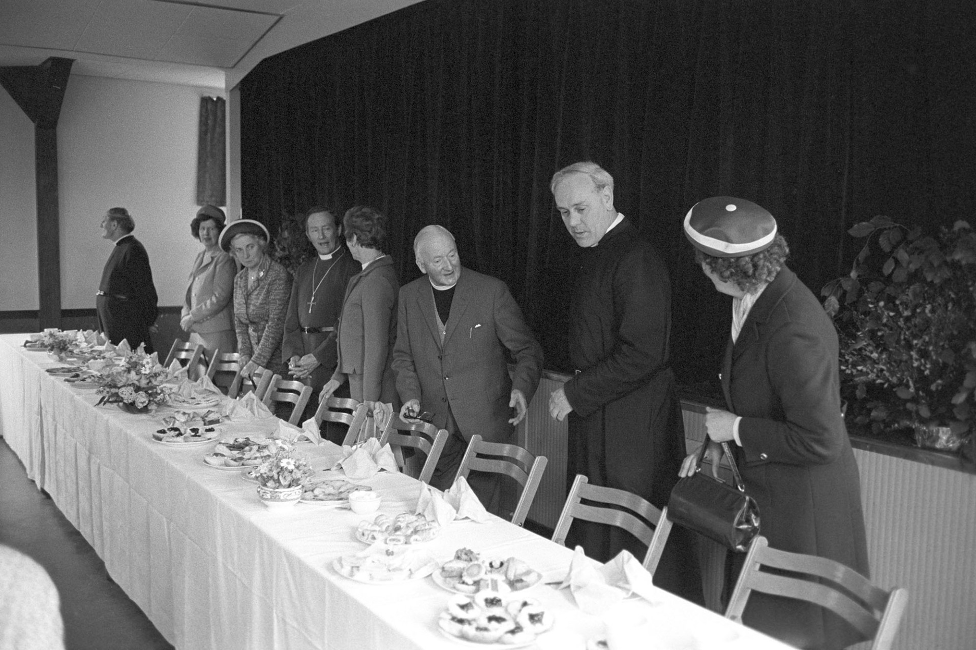 Mothers Union after church tea in the village hall vicars and wives.
[Reverend Moorse, in the centre, and other vicars and their wives waiting to be seated at a table spread with afternoon tea in Dolton Village Hall after the Mother's Union Parade. The table is decorated with small flower arrangements.]