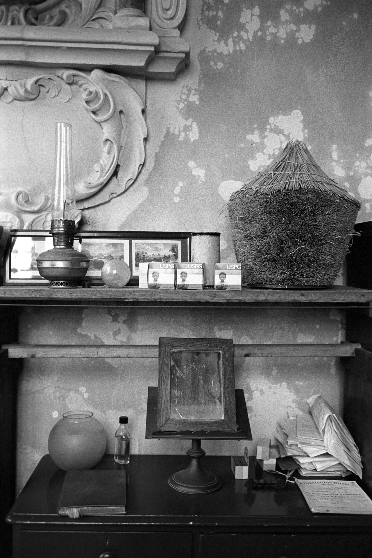 Interior of church vestry, with oil lamp, mirror, and small model rick for Harvest Festivals.
[Desk and shelf in Atherington Church vestry with an oil lamp, pictures, mirror, papers and model thatched wheat rick, amongst other items. Plasterwork can be seen on the wall behind, next to flaking paintwork.]