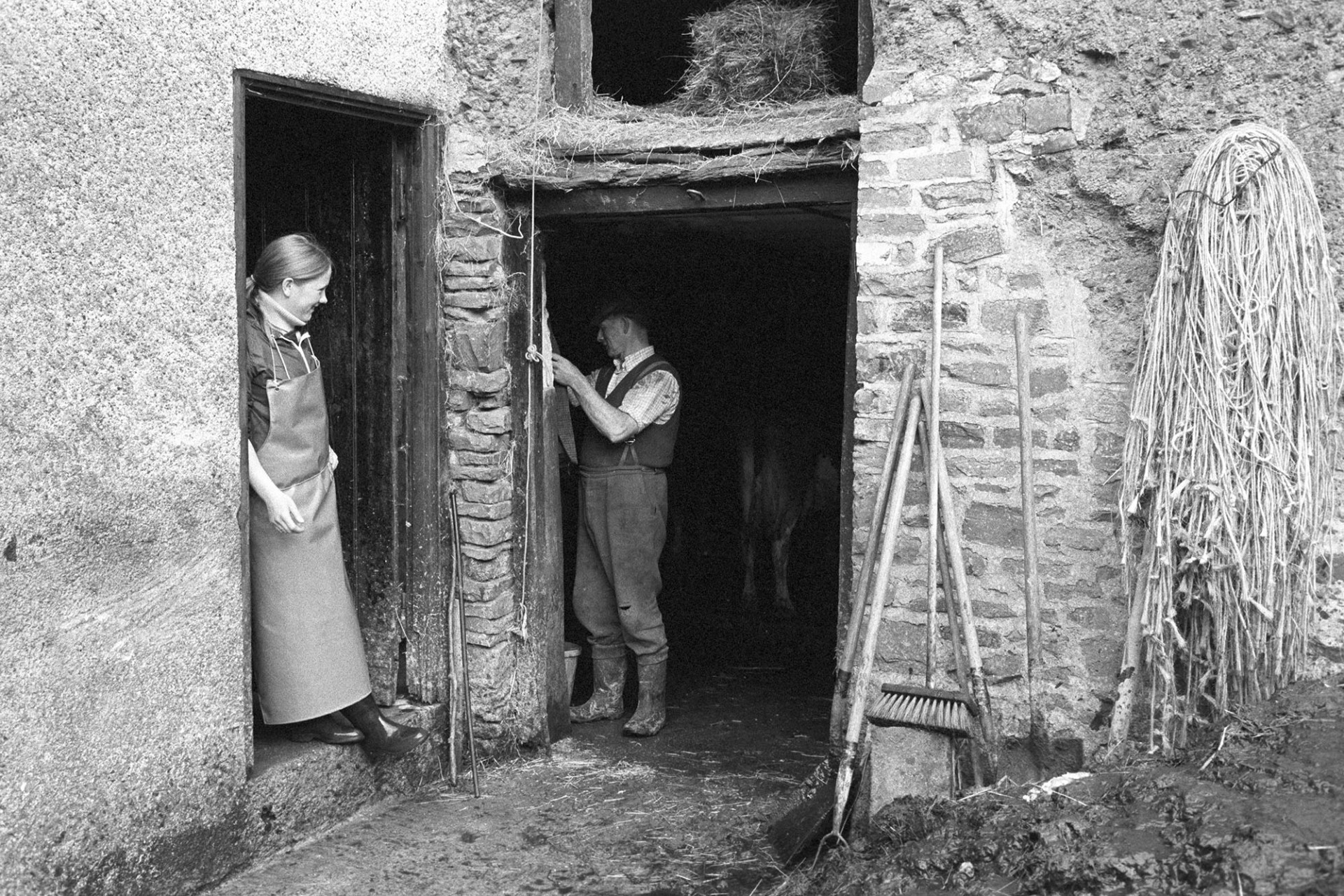 Farmer and daughter at barn door before milking.
[John Woolacott working in a barn and talking to his daughter who is leaning in a doorway. Farm tools are lent against the barn wall next to ropes hanging up. A hay bale can be seen in the barn loft.]