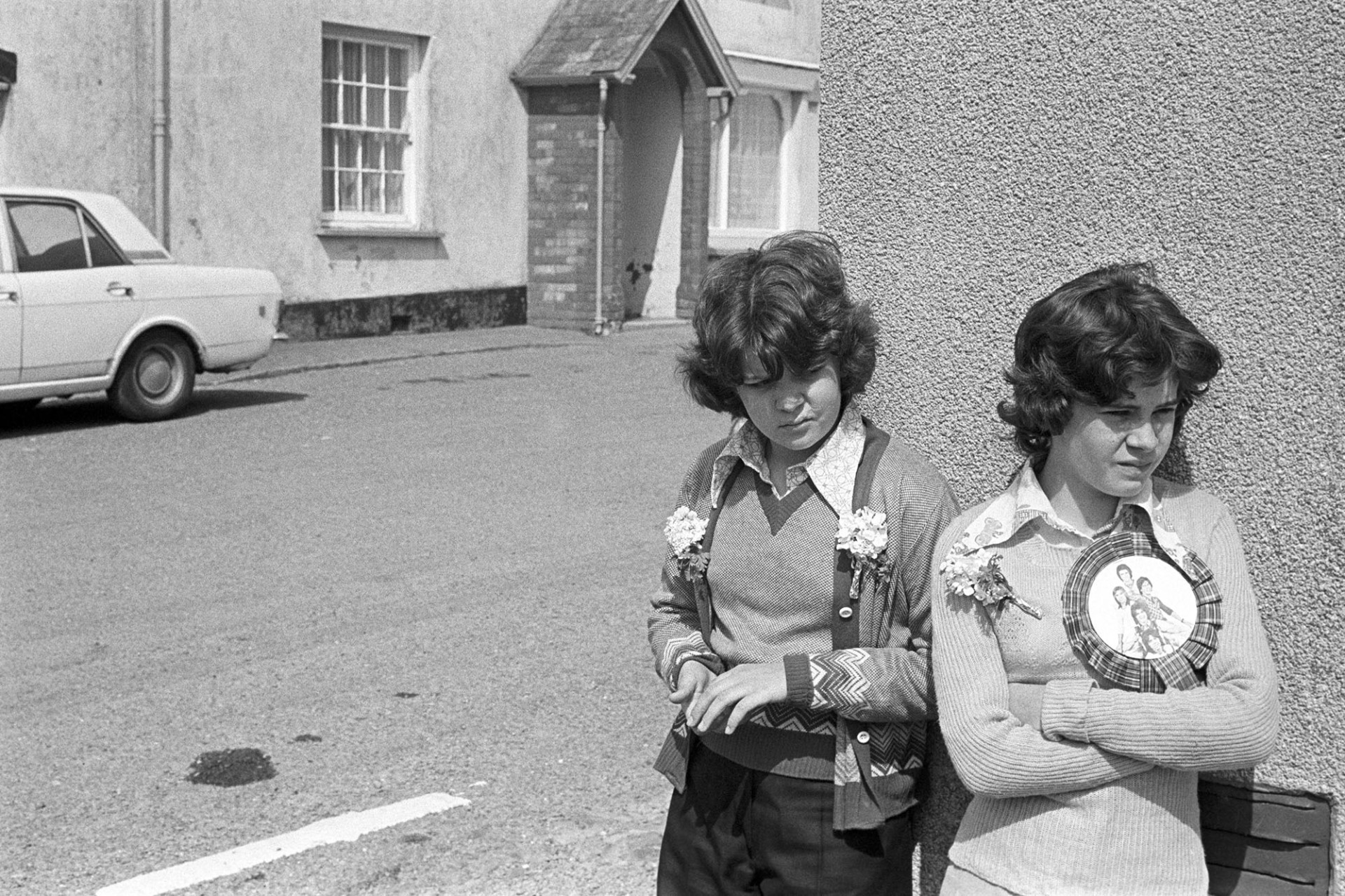 Two girls on street corner in village.
[Two young women leaning against a wall in a street in High Bickington. Both girls are wearing buttonholes and one is wearing a large badge, possibly with the picture of a band on the front.]