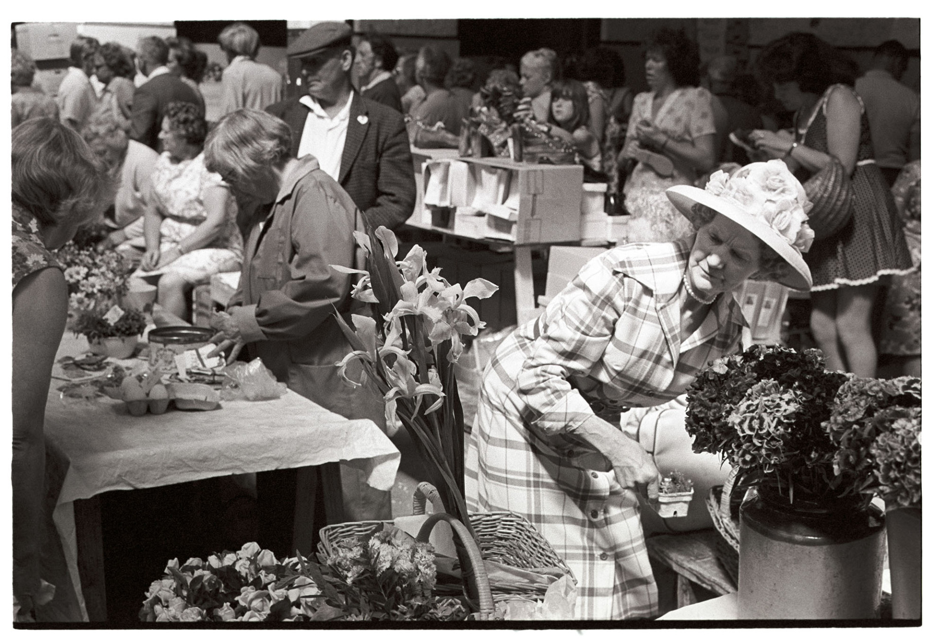 Flower stall at pannier market, woman in flowery hat.
[People shopping in Barnstaple Pannier Market. A woman wearing a hat with flowers is running a flower stall at the market.]