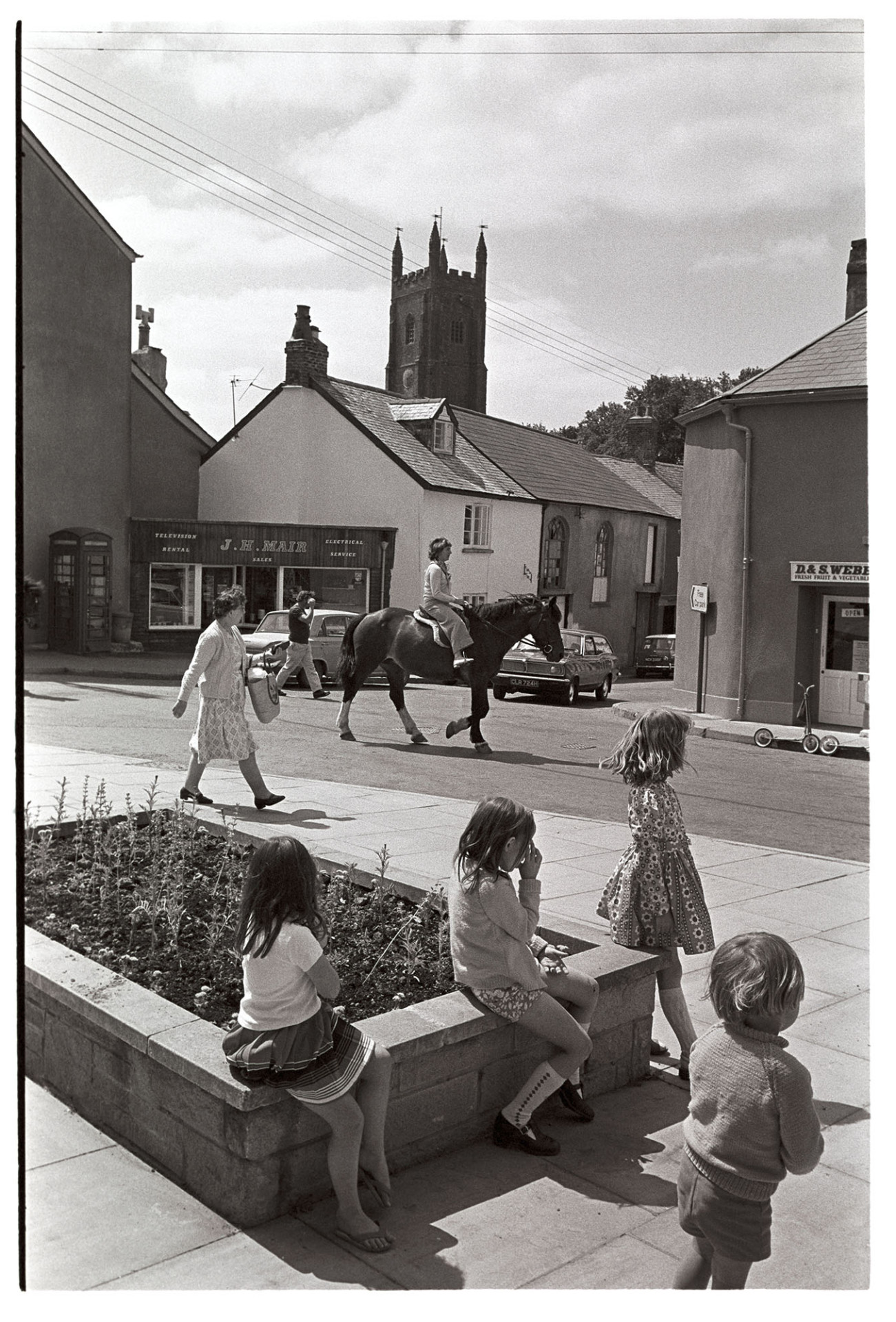 Children sitting in town.
[Children sitting on a raised flower bed in Chulmleigh town centre, as a woman rides past on horseback. The shop front of J H Mair, television rental and electrical service, and the church tower is visible in the background]
