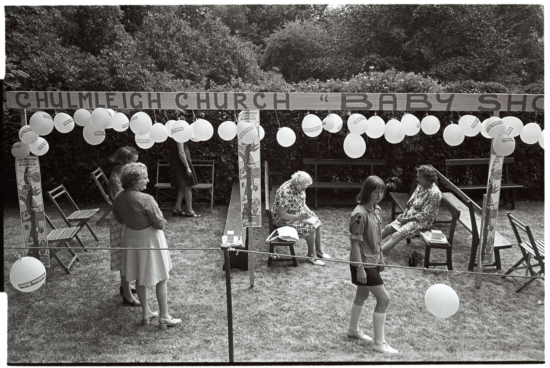 Church Garden Party, arena for baby show with balloons.
[Six women in the balloon decorated area for the baby show at the Chulmleigh Church Garden Party at Chulmleigh Vicarage. Some of the women are sat on benches.]