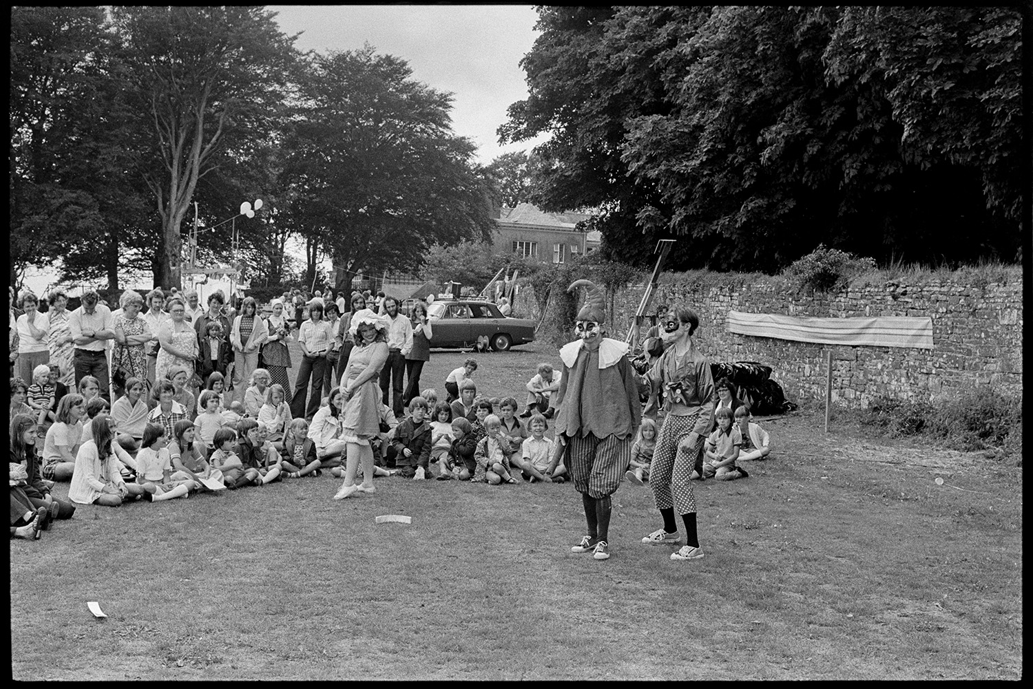 Performance of play at village revel. 
[A group of children sat on the grass wth adults stood behind watching an open air play at the Beaford Revel on the village green. The three actors performing are dressed in costumes.]