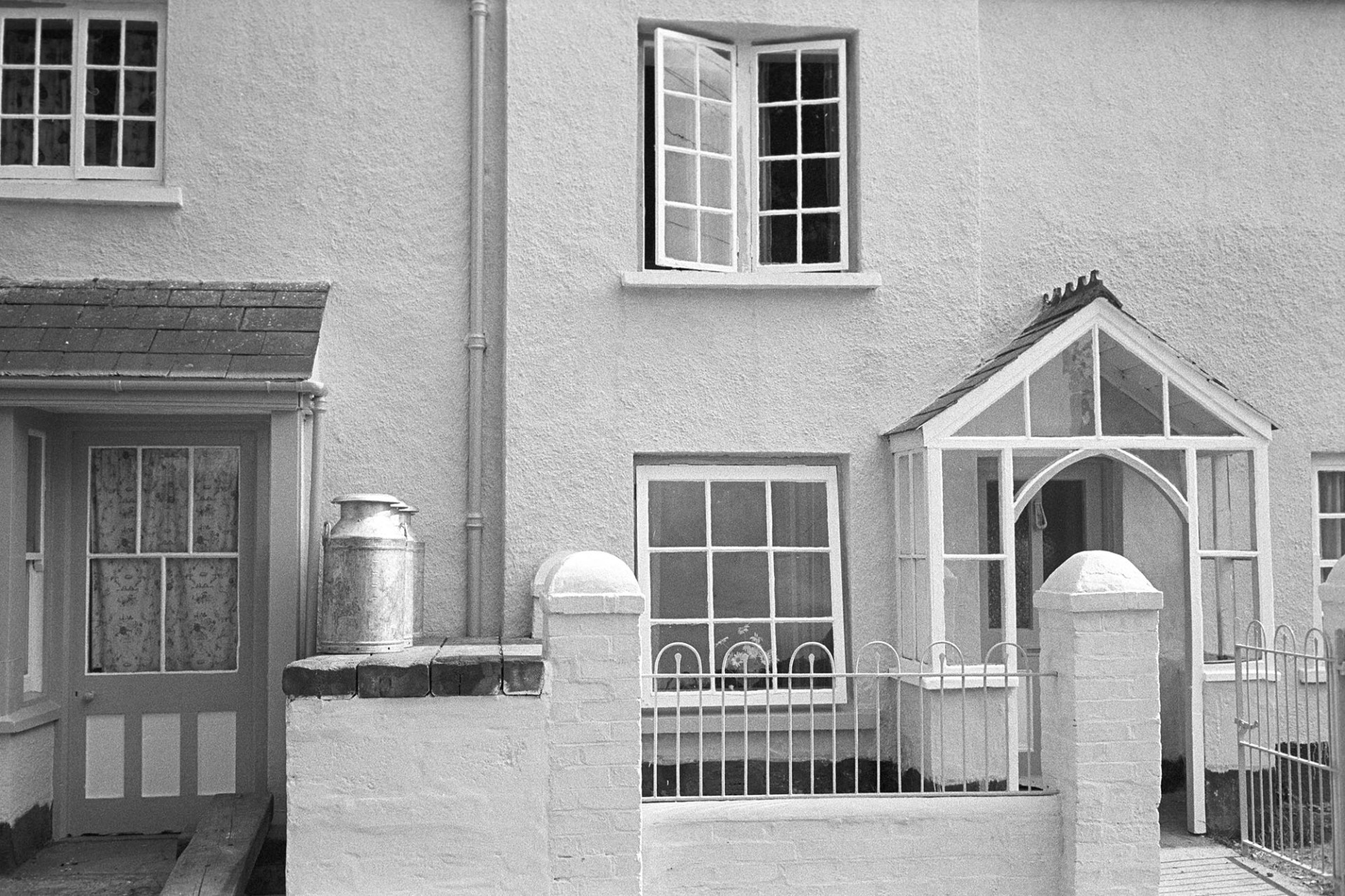 Front of house with milk stand and milk churns.
[Three milk churns on a milk stand outside a house in East Street, Sheepwash.]