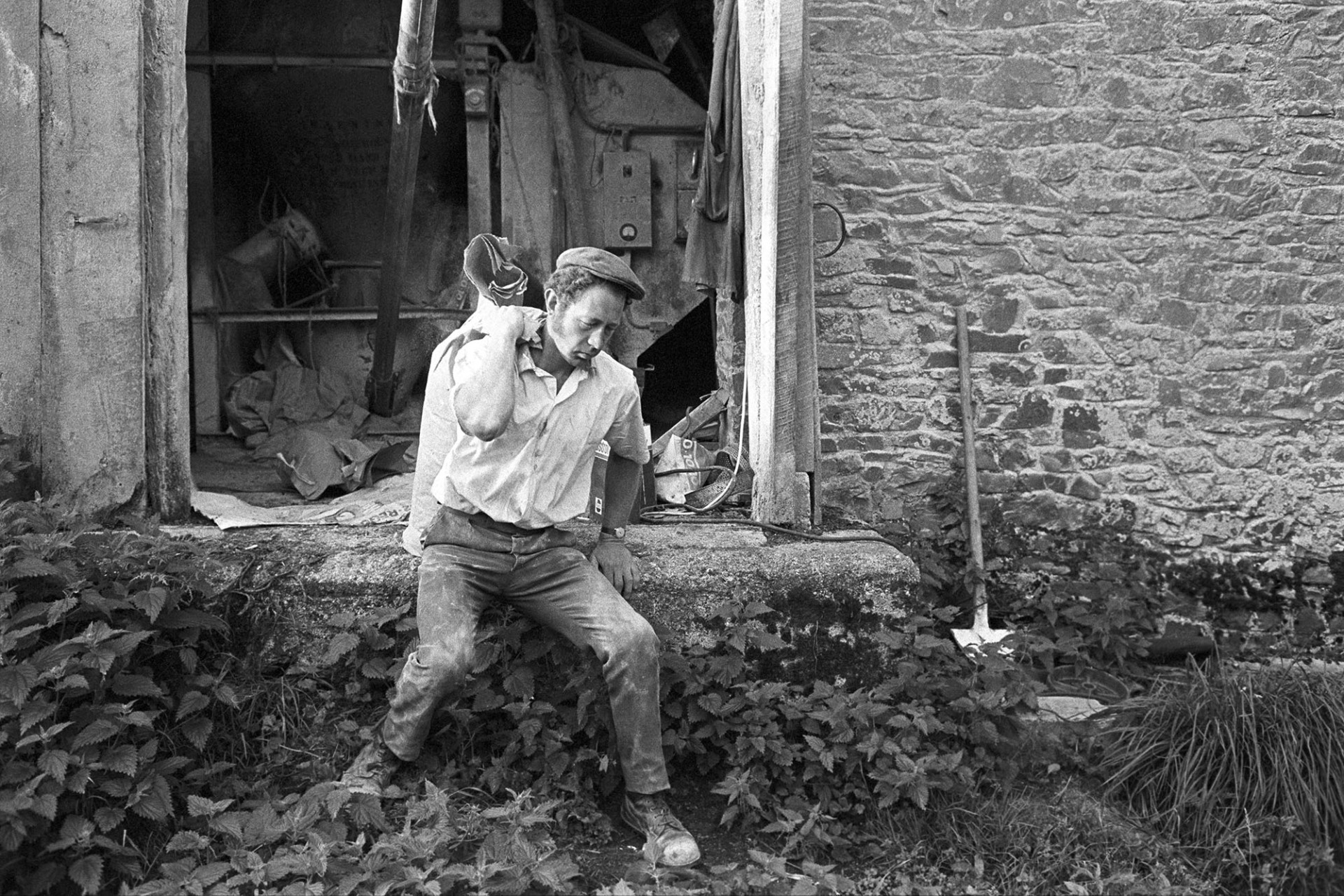 Farmer lifting sack at barn entrance.
[Mr Pengelly lifting a sack onto his back at a barn entrance at Woolleigh Barton, Beaford. The entrance is surrounded by stinging nettles and a shovel is propped up against the barn wall.]