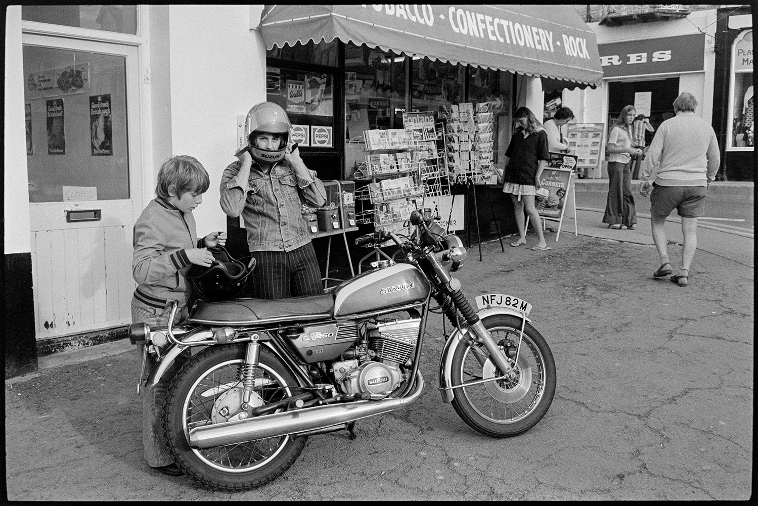 Motorbike and rider at seaside. 
[A motocyclist and child putting on helmets next to their motorbike which is parked outside a giftshop at Westward Ho! A woman is looking at postcards displayed outside the giftshop.]