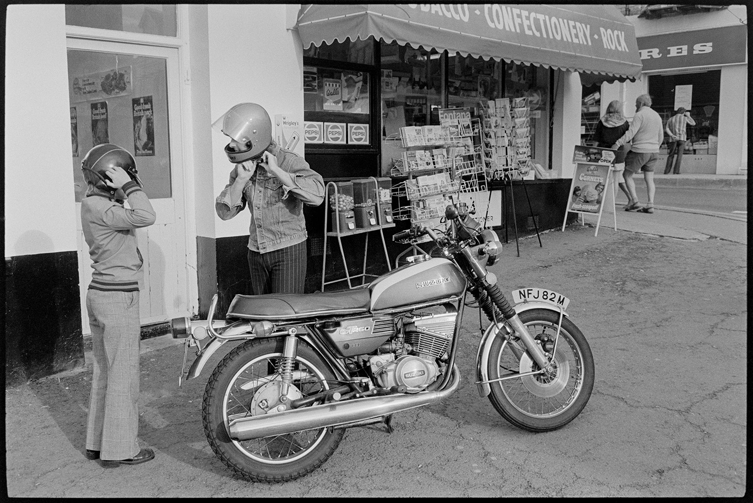 Motorbike and rider at seasid. 
[A motocyclist and child putting on helmets next to their motorbike which is parked outside a giftshop at Westward Ho! Postcards are sweets are displayed outside the giftshop.]