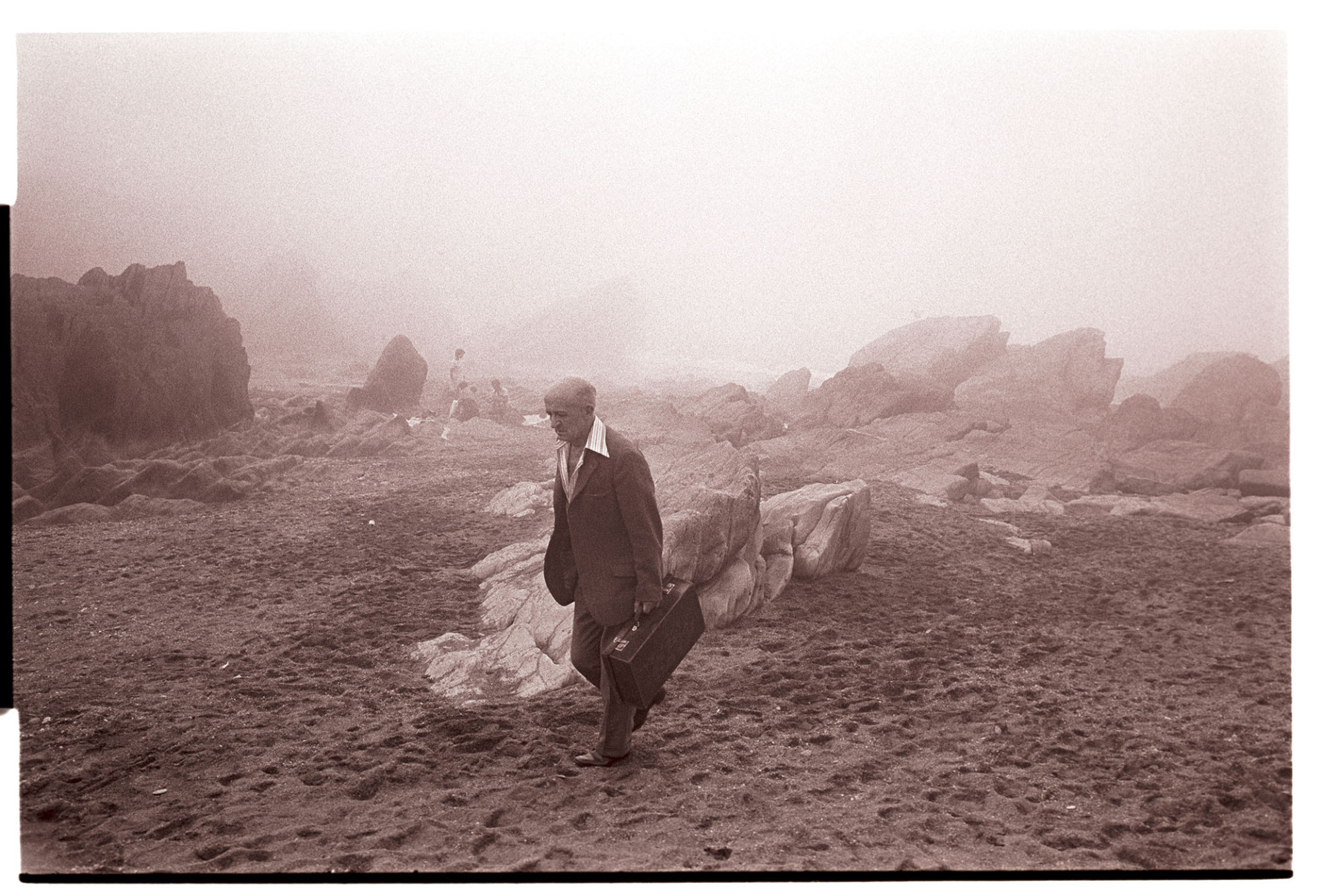 Holidaymakers on rocky shore in thick sea mist.
[A man wearing a suit and carrying a small suitcase walking along Ilfracombe beach in the mist. Holidaymakers are visible in the background by rocks.]