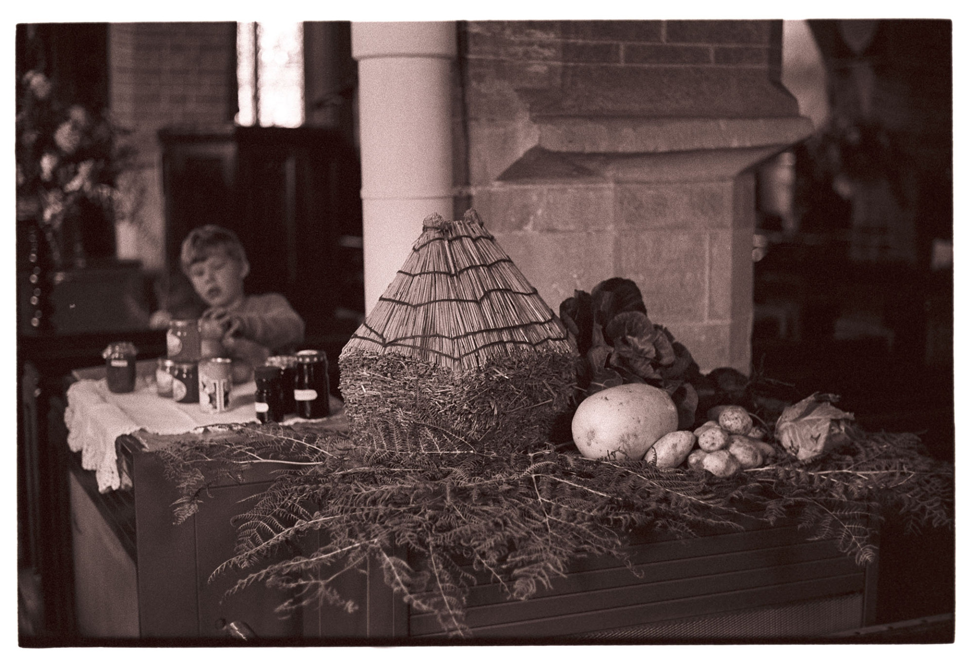 Display of produce in church Harvest Festival, with small thatched rick.
[A harvest festival display in Dolton church with vegetables, ferns and a  model thatched rick. A boy is stacking jars of jam or marmalade on a table in the background.]
