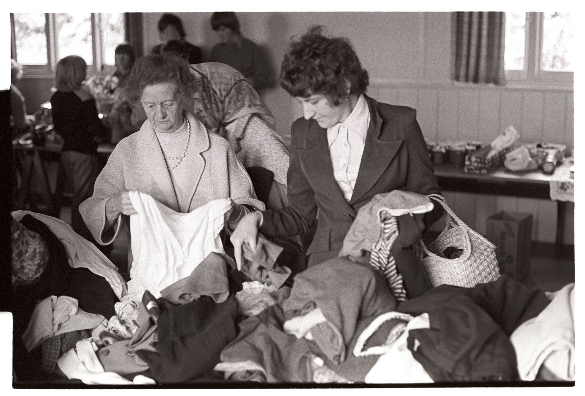 Women looking at clothes at jumble sale.<br /> [Two women looking at clothes in a jumble sale at Harracott. Other stalls are visible in the background.]