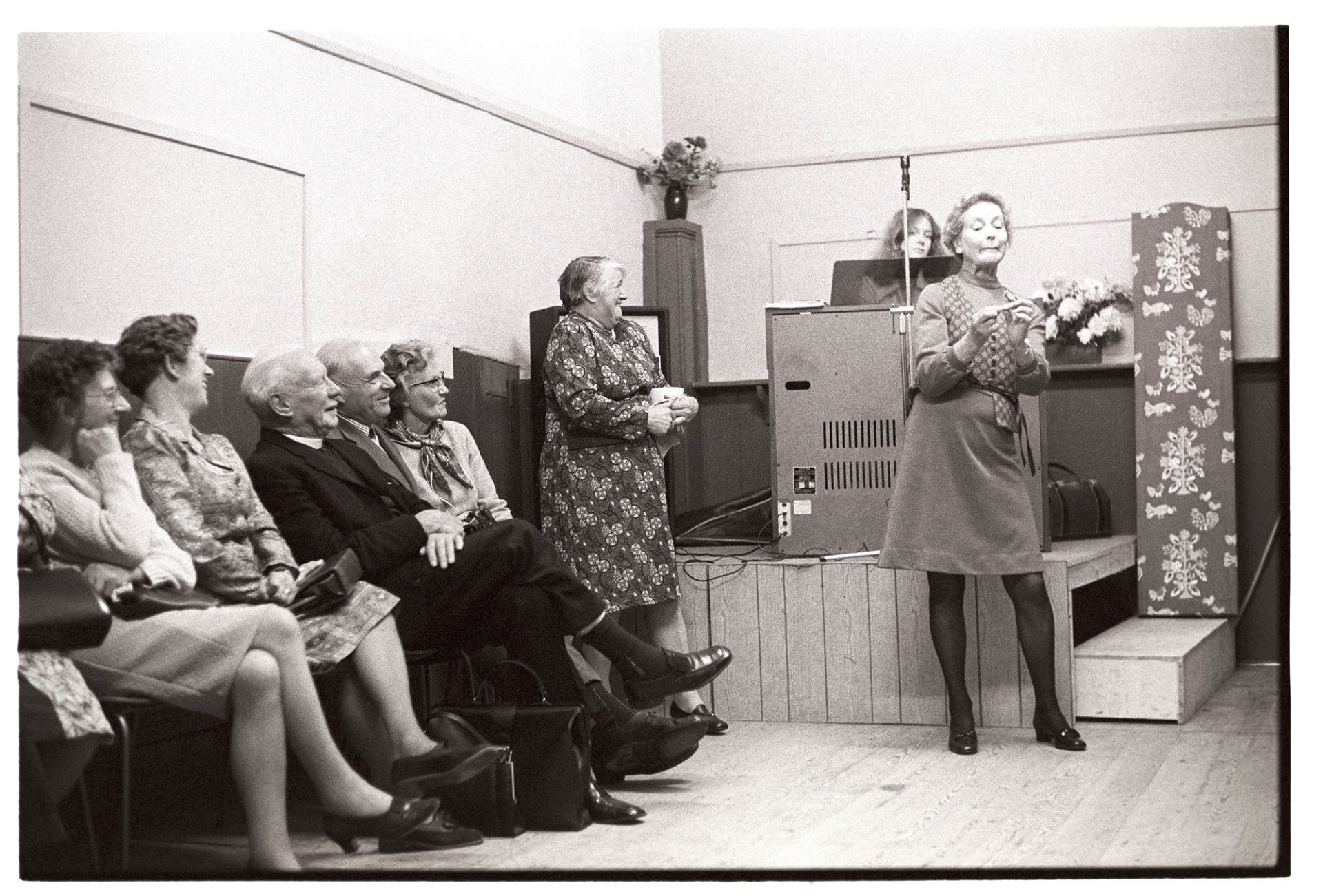 Entertainment at Harvest Supper in village hall.
[Reverend Wallington and other people watching Mrs Wallington perform a mime to entertain people at the Harvest Supper at Roborough Village Hall. A woman is stood up laughing by a small stage.]