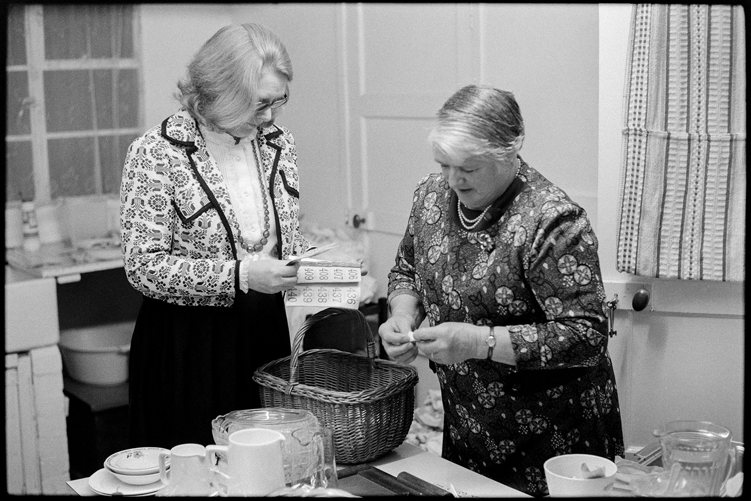 Women preparing food for harvest supper. 
[Mrs Rickman and Mrs Isaacs ripping up raffle tickets and putting them in wicker basket at the harvest supper in Roborough Village Hall. They are working on a table with jugs and plates.]