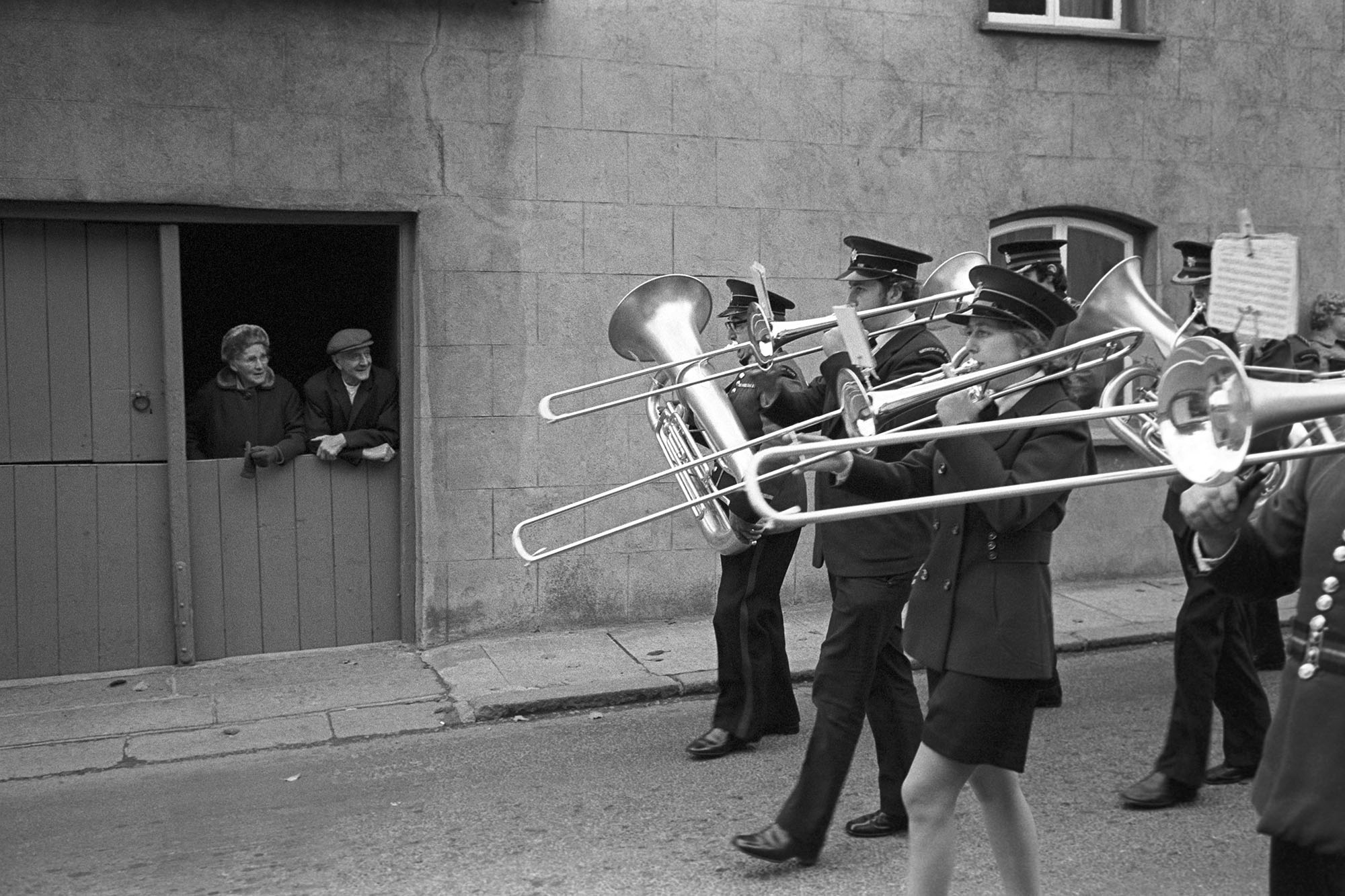 Couple watching band passing their door. Parade.
[A man and woman leaning on a stable door watching a brass band parade past at Hatherleigh Carnival.]