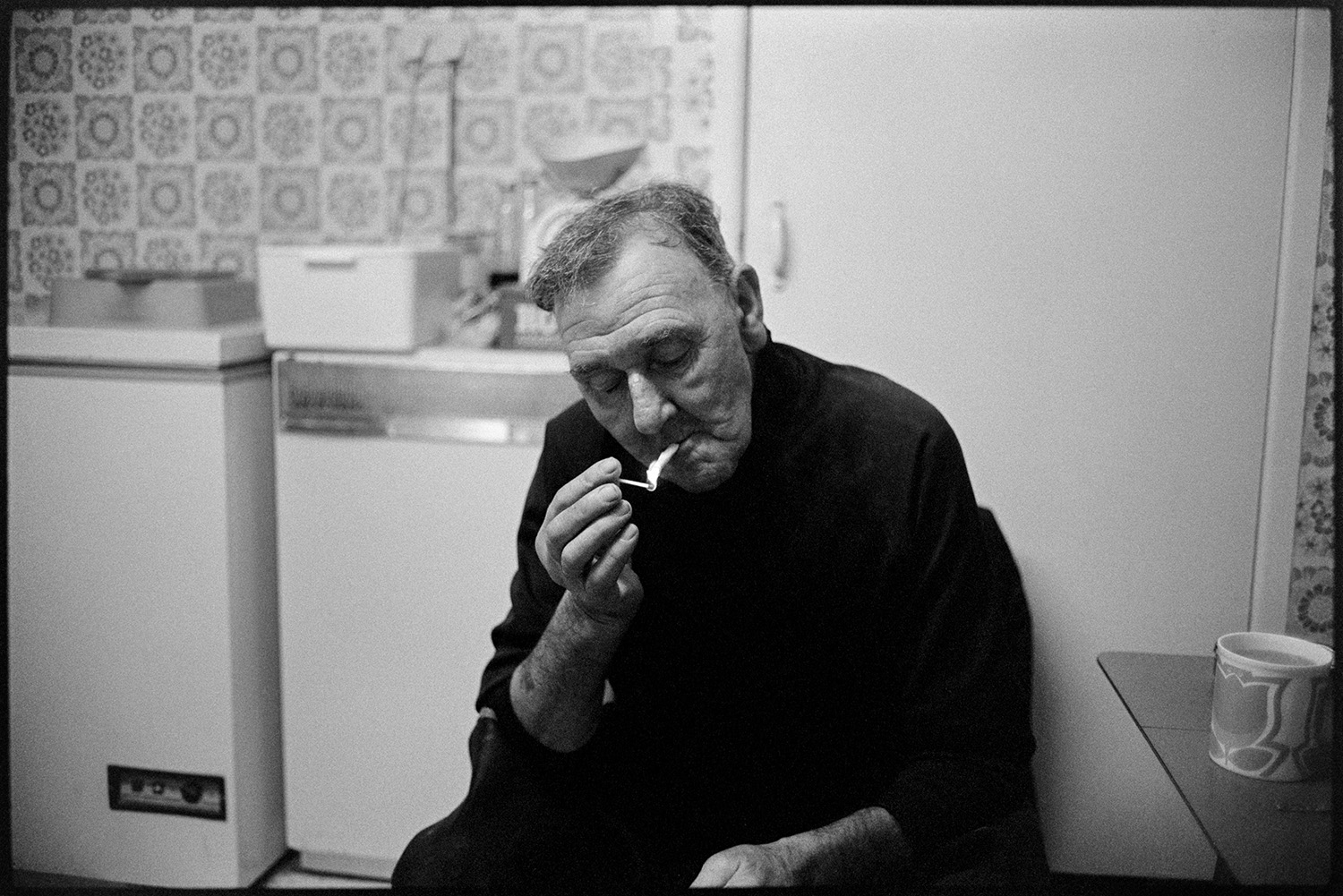 Couple preparing laver in kitchen. 
[Tim Schiller sat in his kitchen lighting a cigarette with a match. A fridge or freezer can be seen in the background and a cup of tea is on the table next to him.]