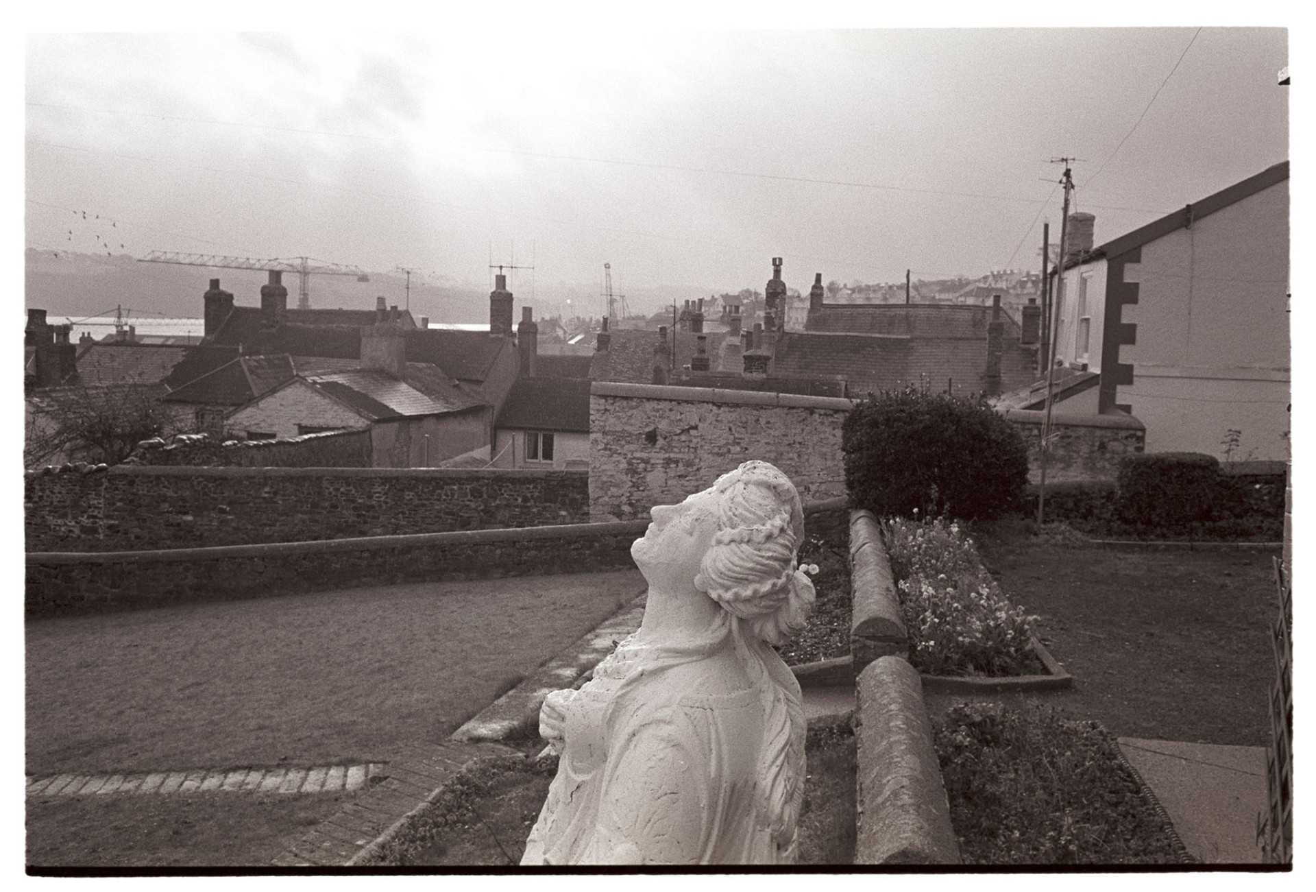 Old ship's figurehead in garden.
[An old ship's figurehead in a garden in Appledore. Houses and rooftops can be seen in the background.]