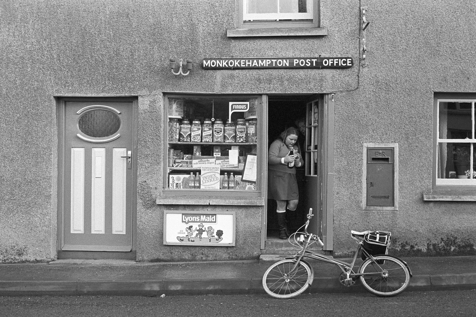 Girl leaving sweetshop to cycle home with sweets!
[A girl leaving Monkokehampton Post Office, with sweets. Her bicycle is leaning against the kerb. A display of sweet jars is visible in the Post Office window and a postbox is to the right of the shop door.]