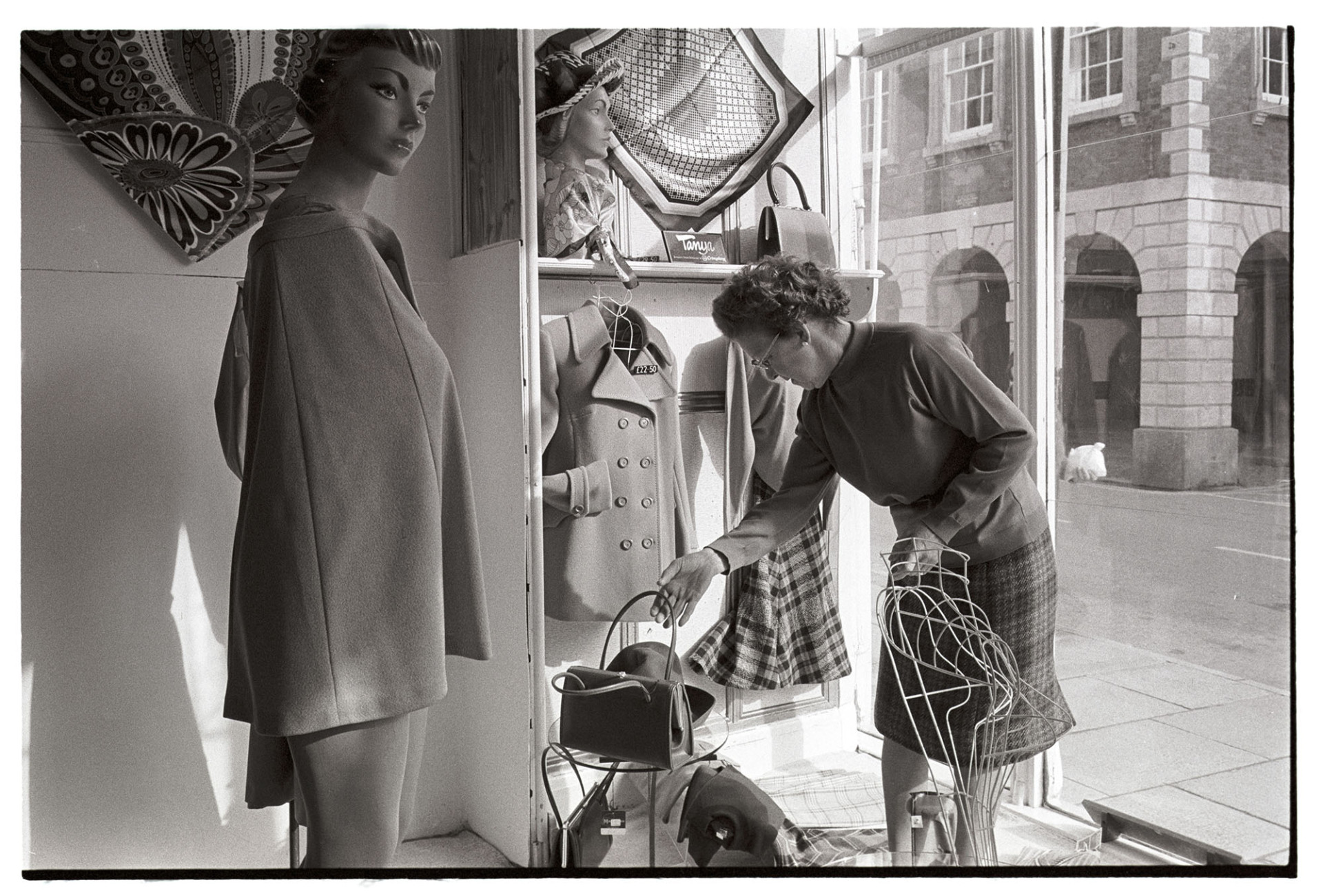 Woman changing window display in women's clothes shop.
[A woman placing a handbag in the window display of a Neals clothes shop in Torrington High Street. Coats and scarves are also on display in the shop front window.]